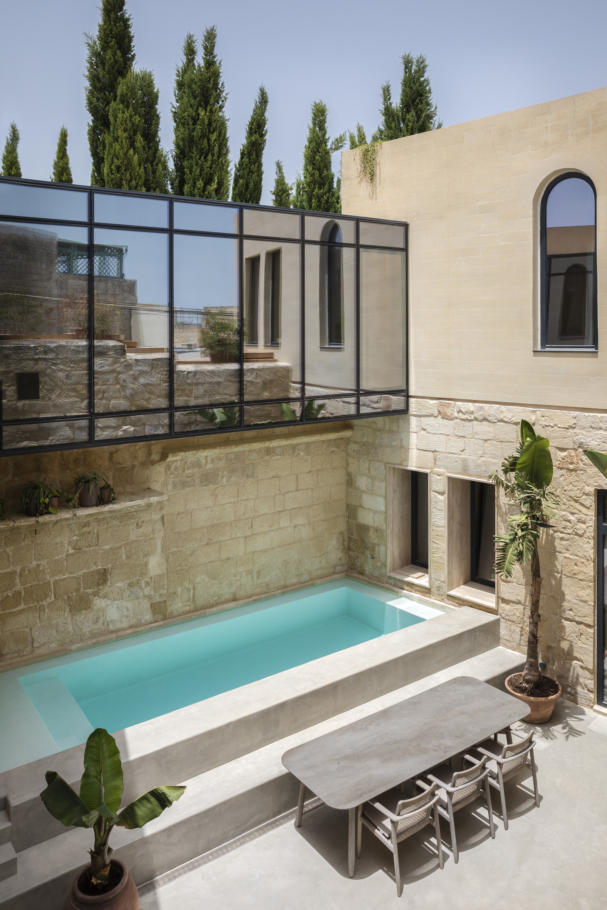 Courtyard pool surrounded by stone walls and black steel glazing with cypresses beyond