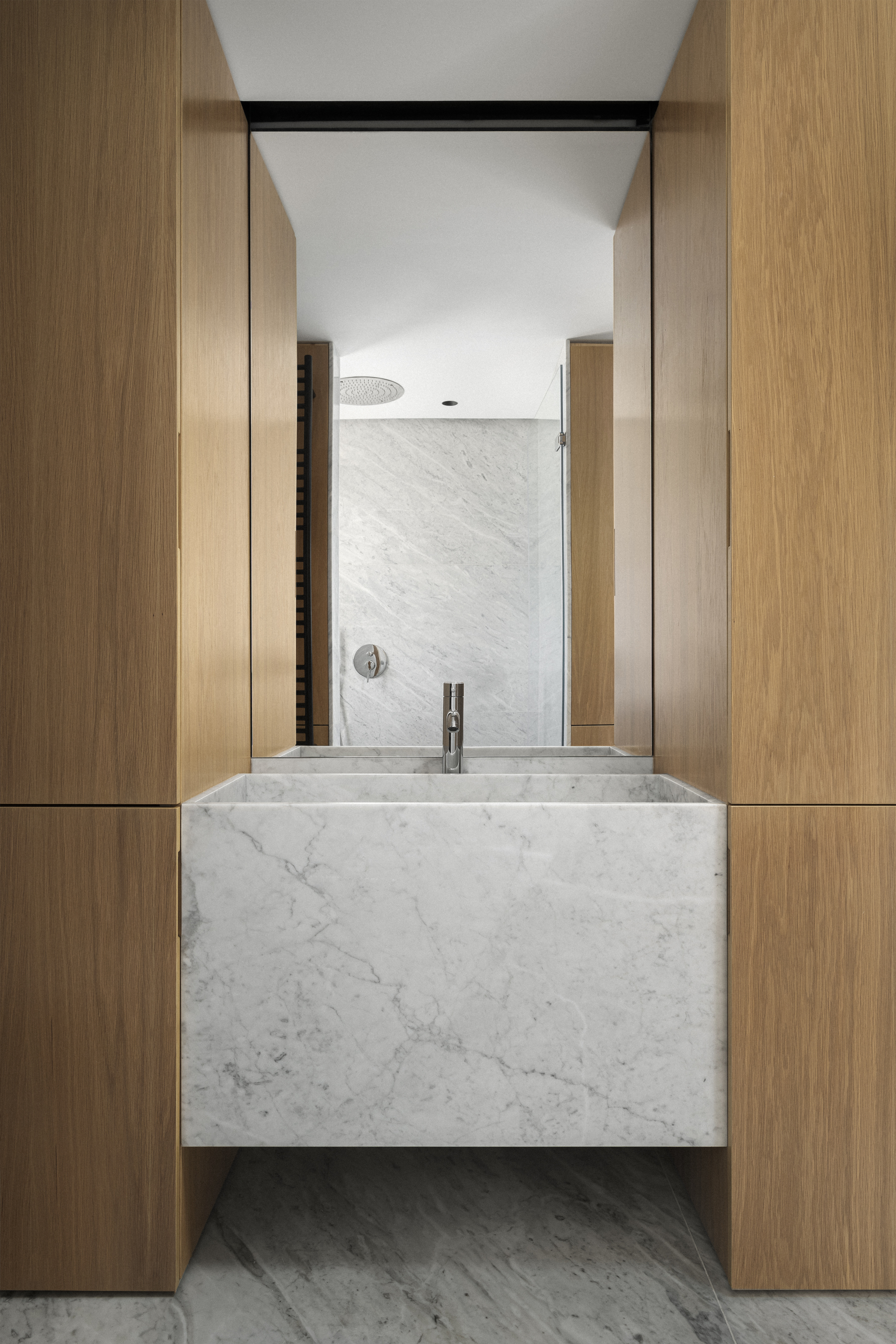 Bathroom vanity in white marble flanked by oak cabinetry with tiled shower beyond