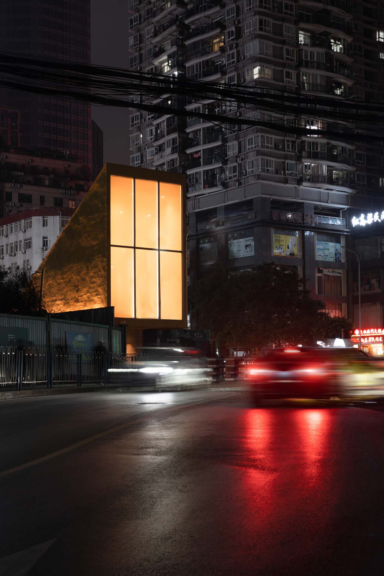 Illuminated glass pavilion with triangular metal facade beside a road with streaking car lights at night