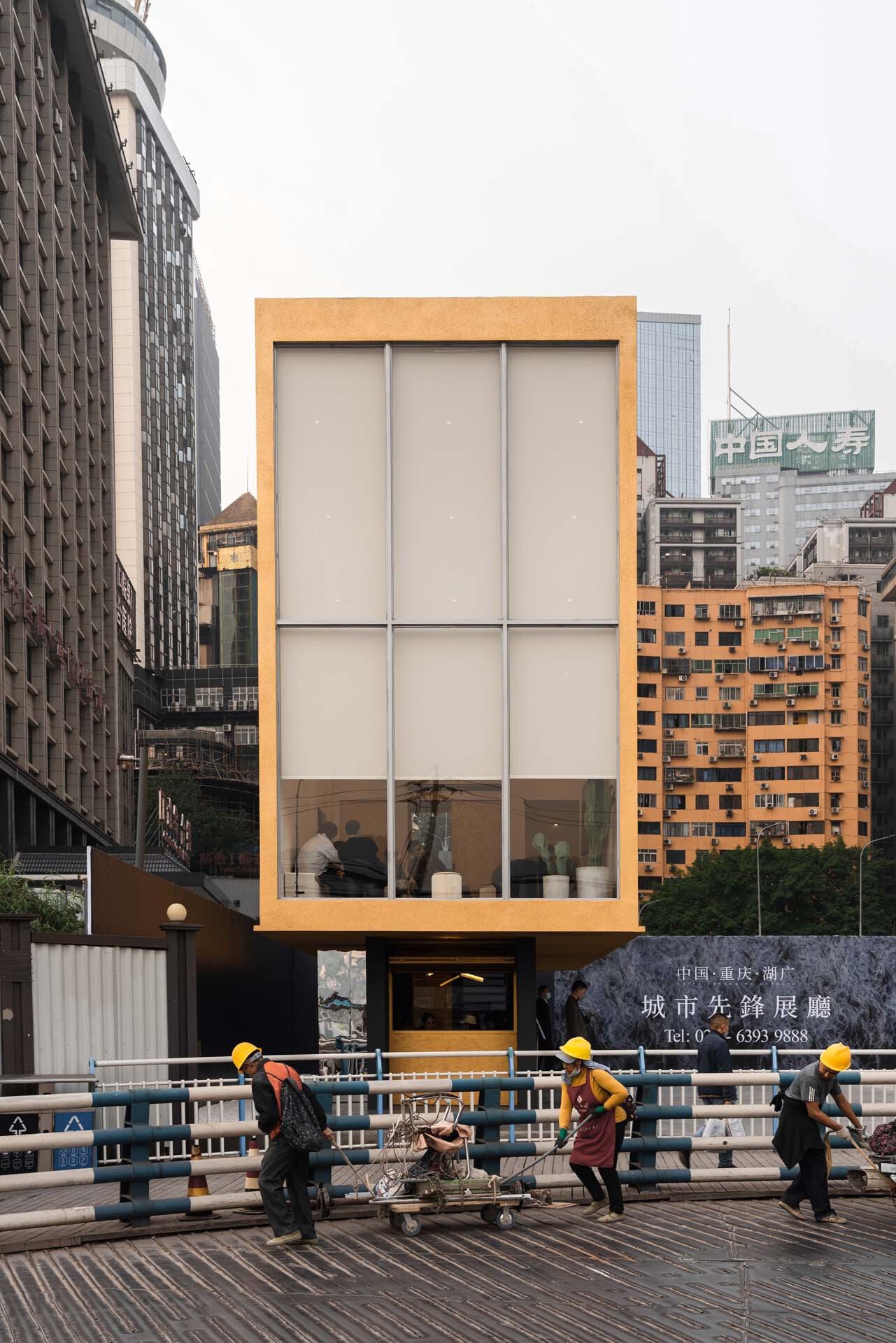 Cantilevered pavilion with yellow frame and translucent panels overlooking workers on the roadway