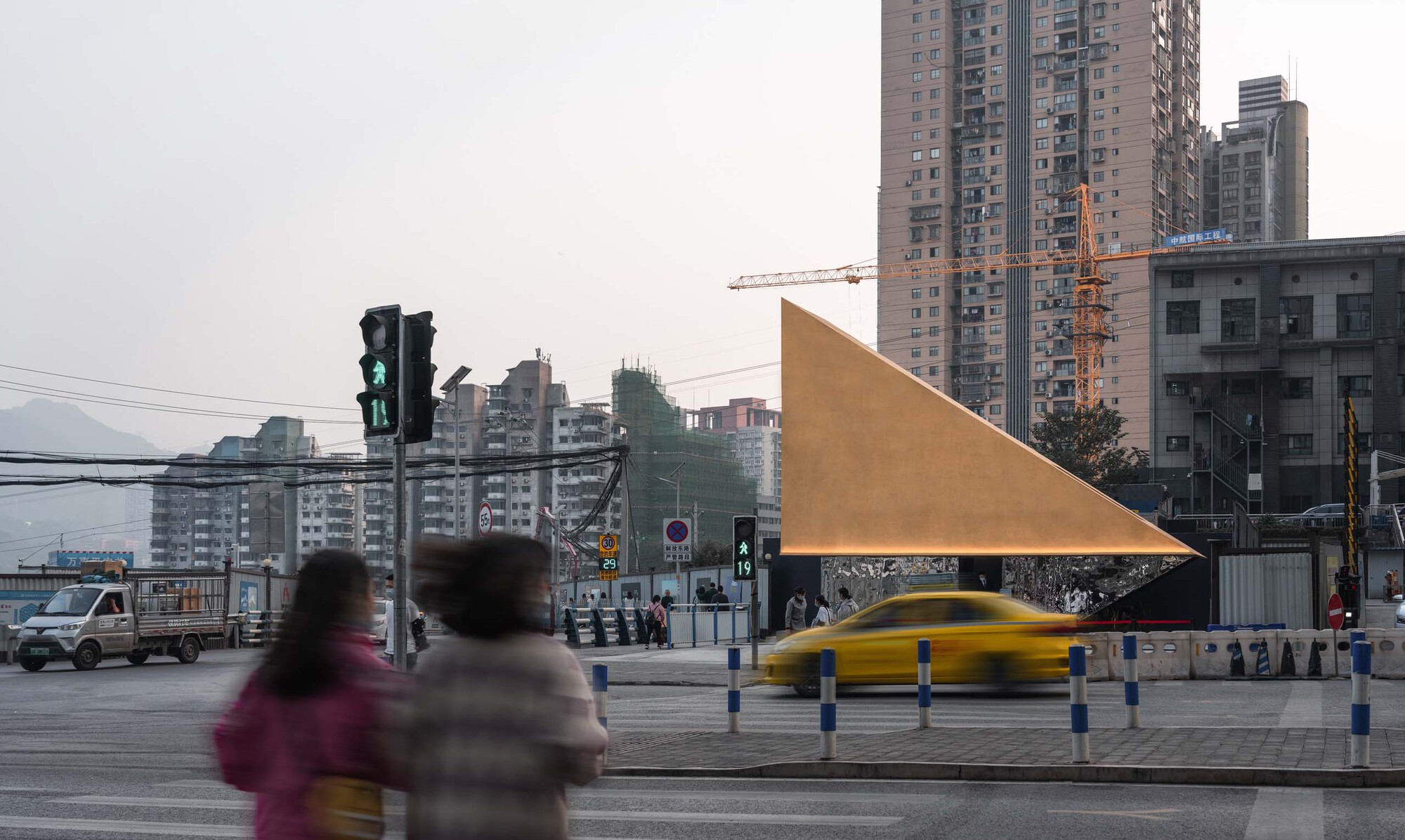 Triangular metal facade viewed from across the intersection with pedestrians and passing taxi