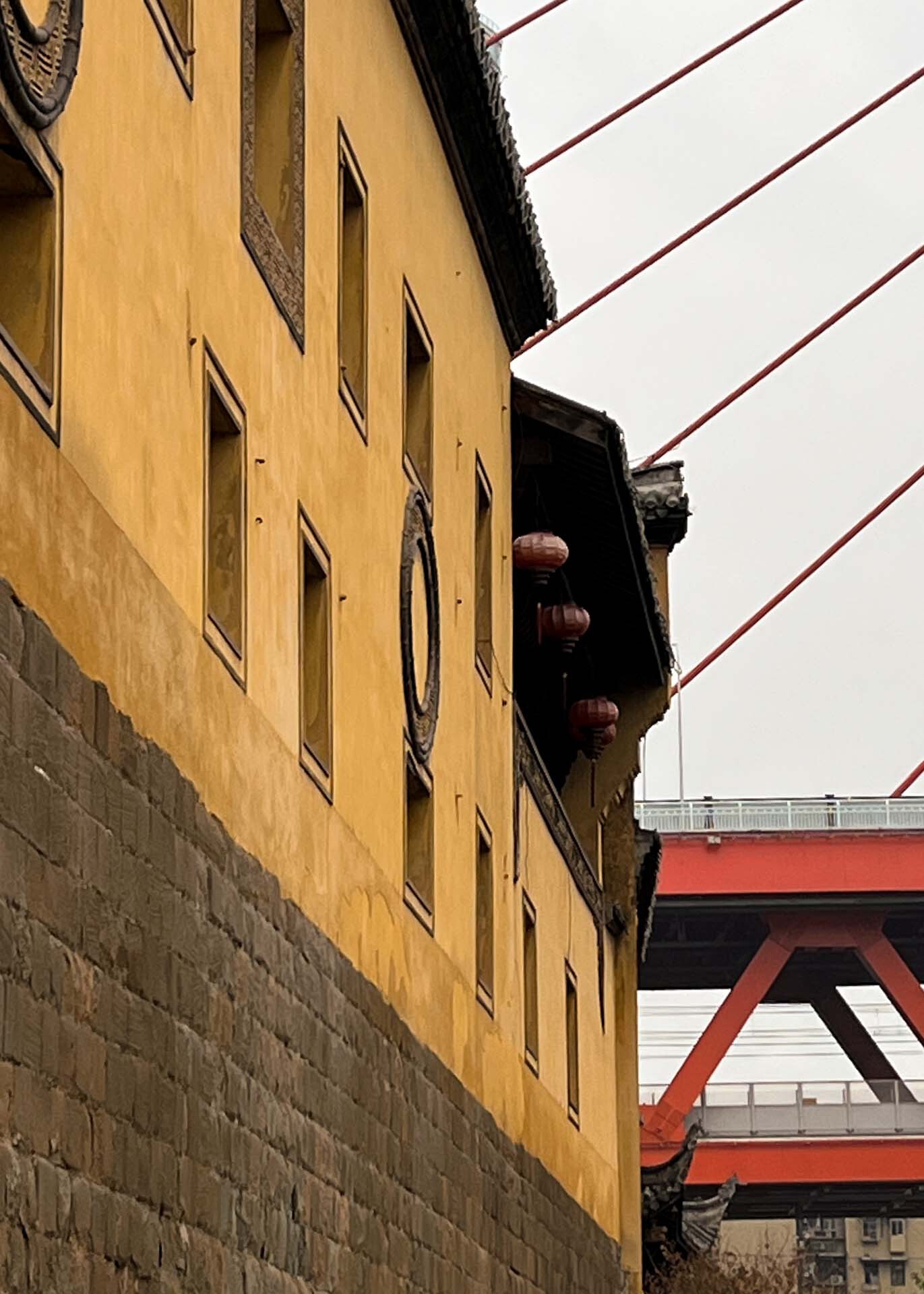 Yellow rendered facade with punched windows beneath the red cables and structure of a bridge