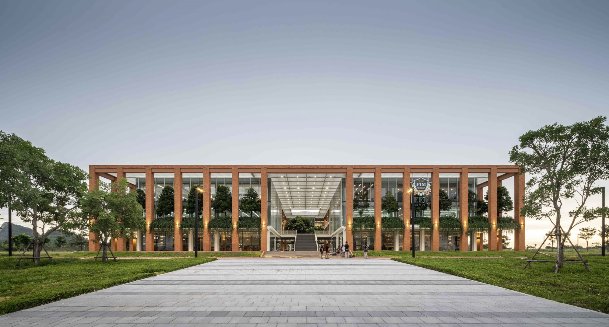 Front elevation of the timber-framed colonnade and glass facade across a paved plaza at dusk