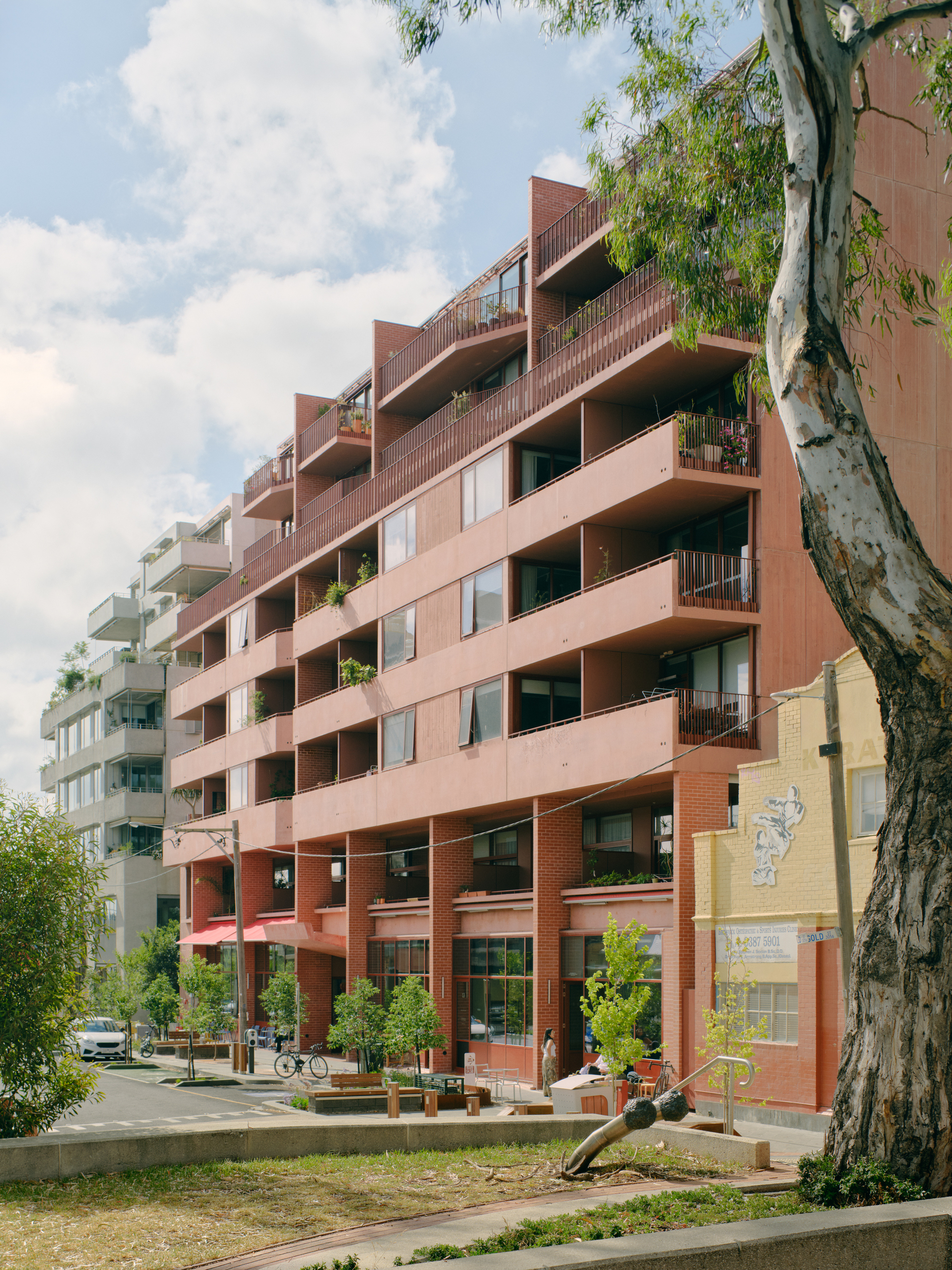 Stepped terracotta facade with planted balconies viewed from tree-lined street under partly cloudy sky