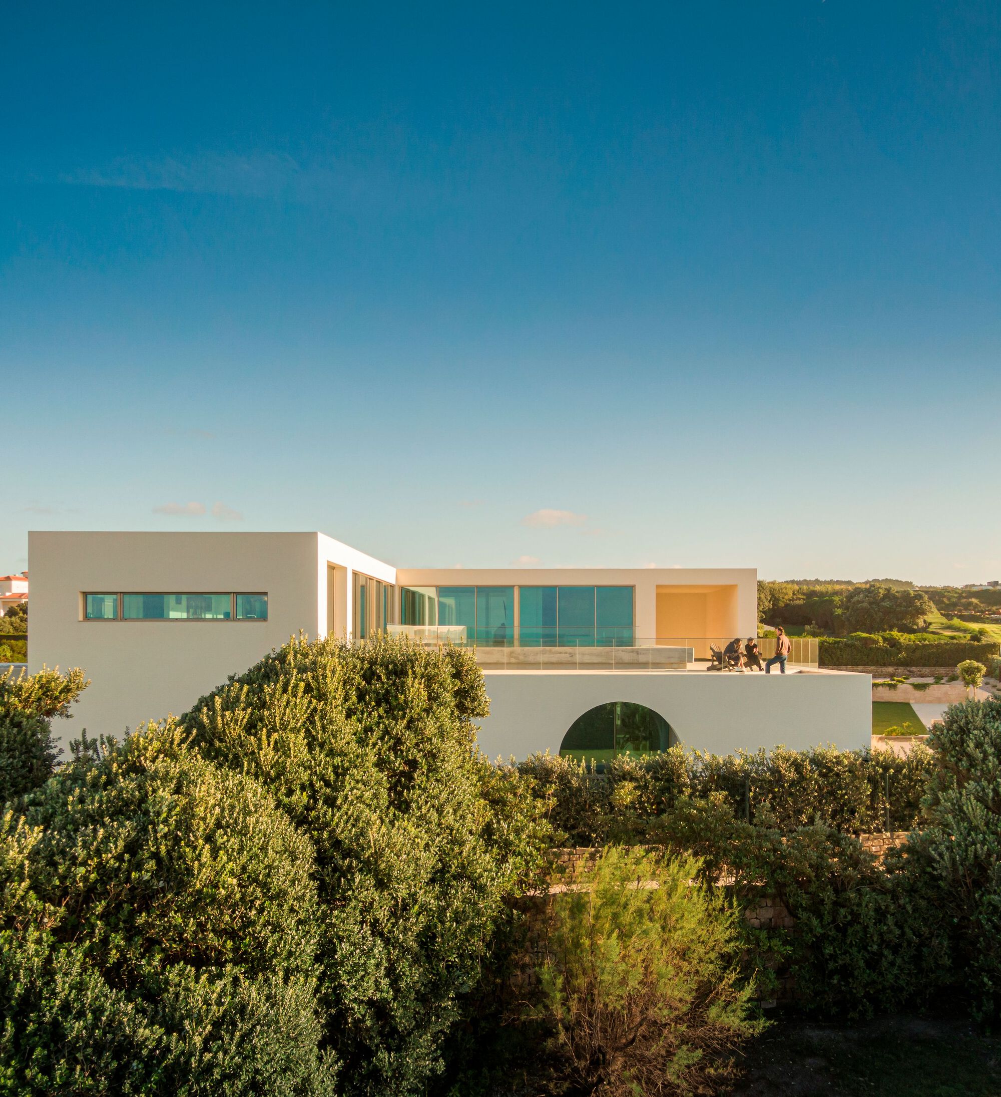Two-story white residence with horizontal ribbon windows and arched opening at base surrounded by dense vegetation