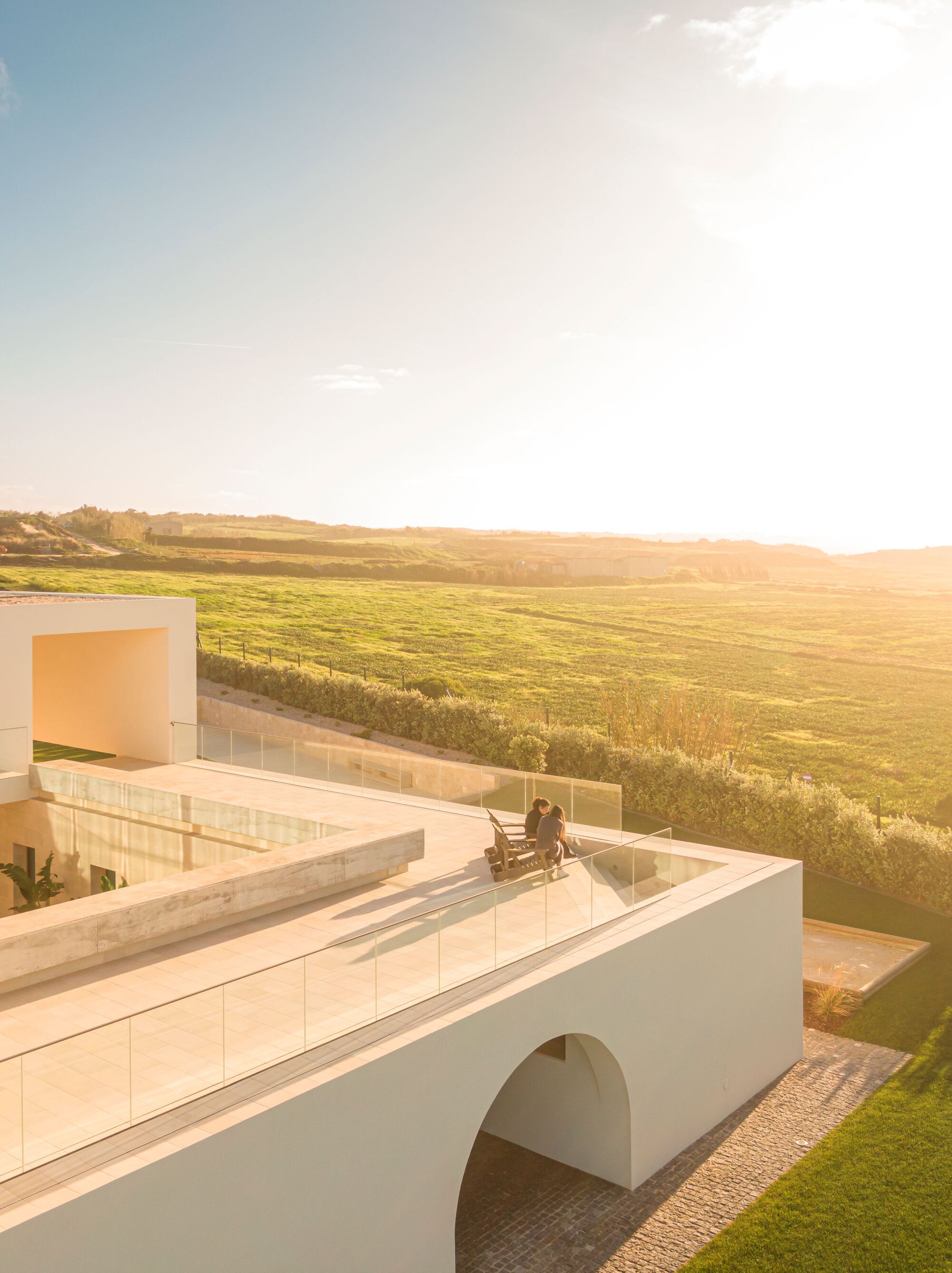 Roof terrace with glass balustrade and person seated in chair overlooking green farmland at golden hour