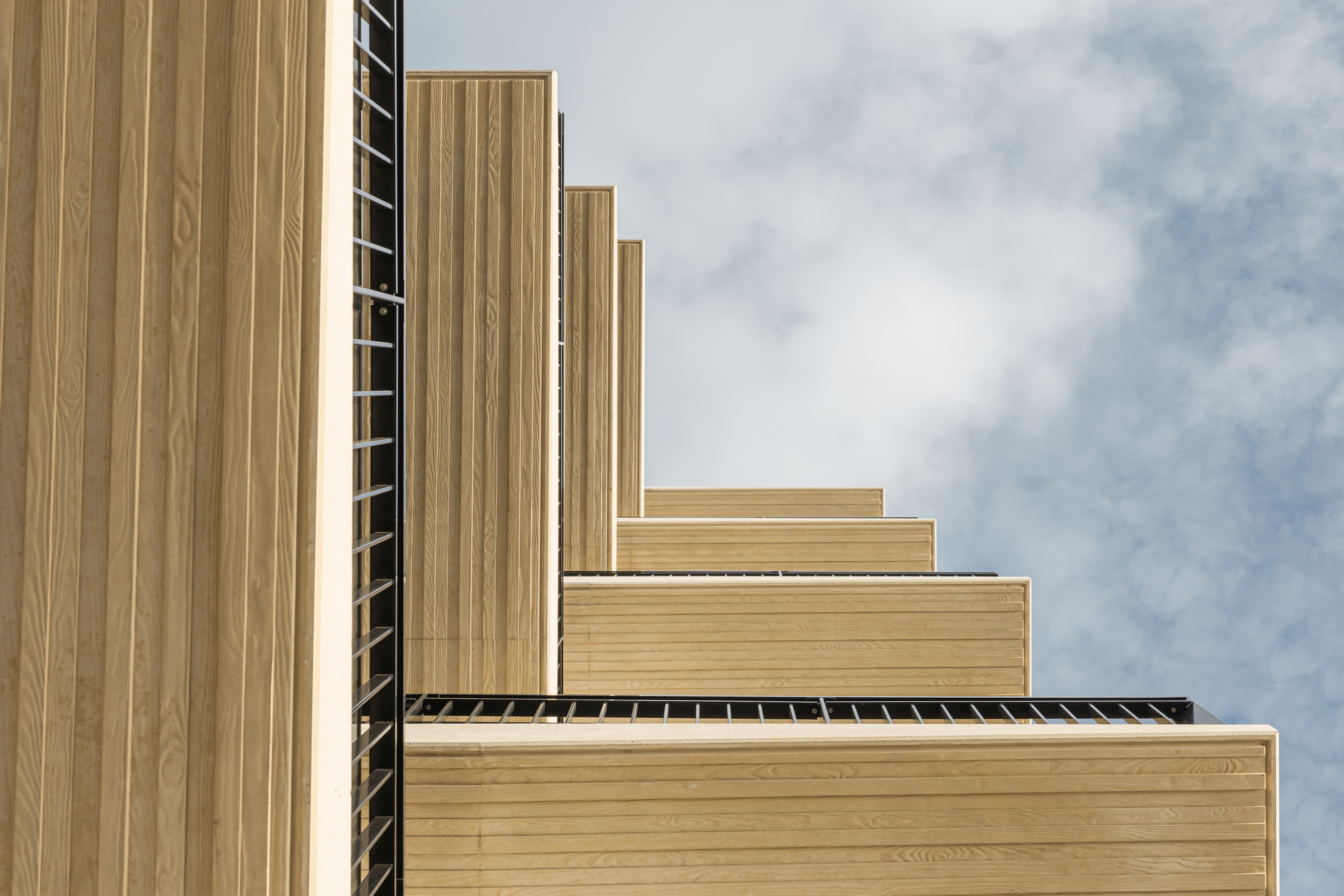 Stepped timber-clad facade volumes with vertical slat screens under a partly cloudy sky