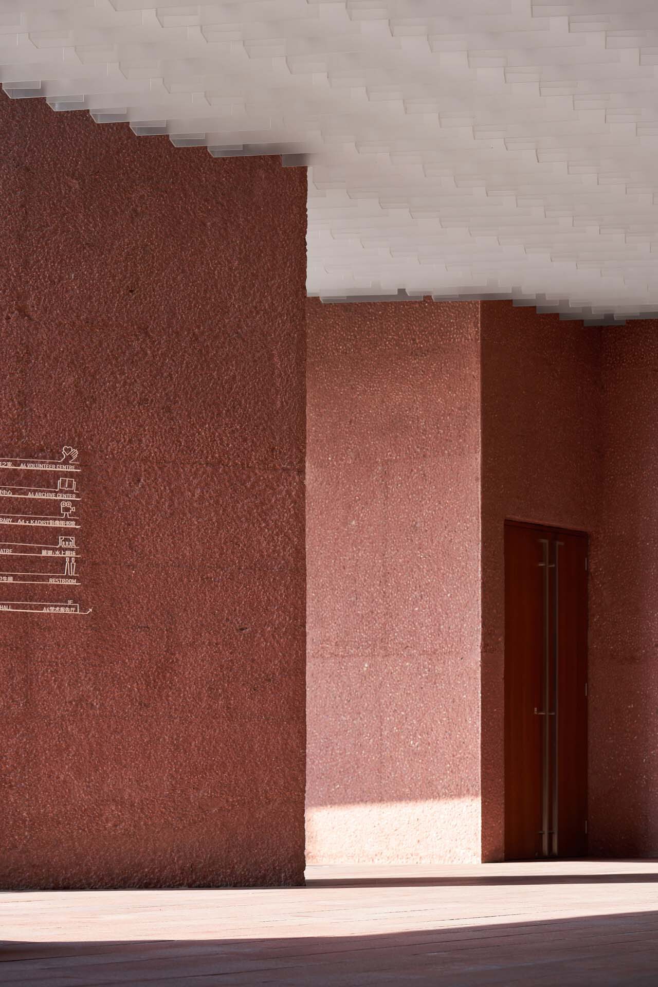 Shaded courtyard corner showing red textured walls and white modular ceiling edge in strong sunlight