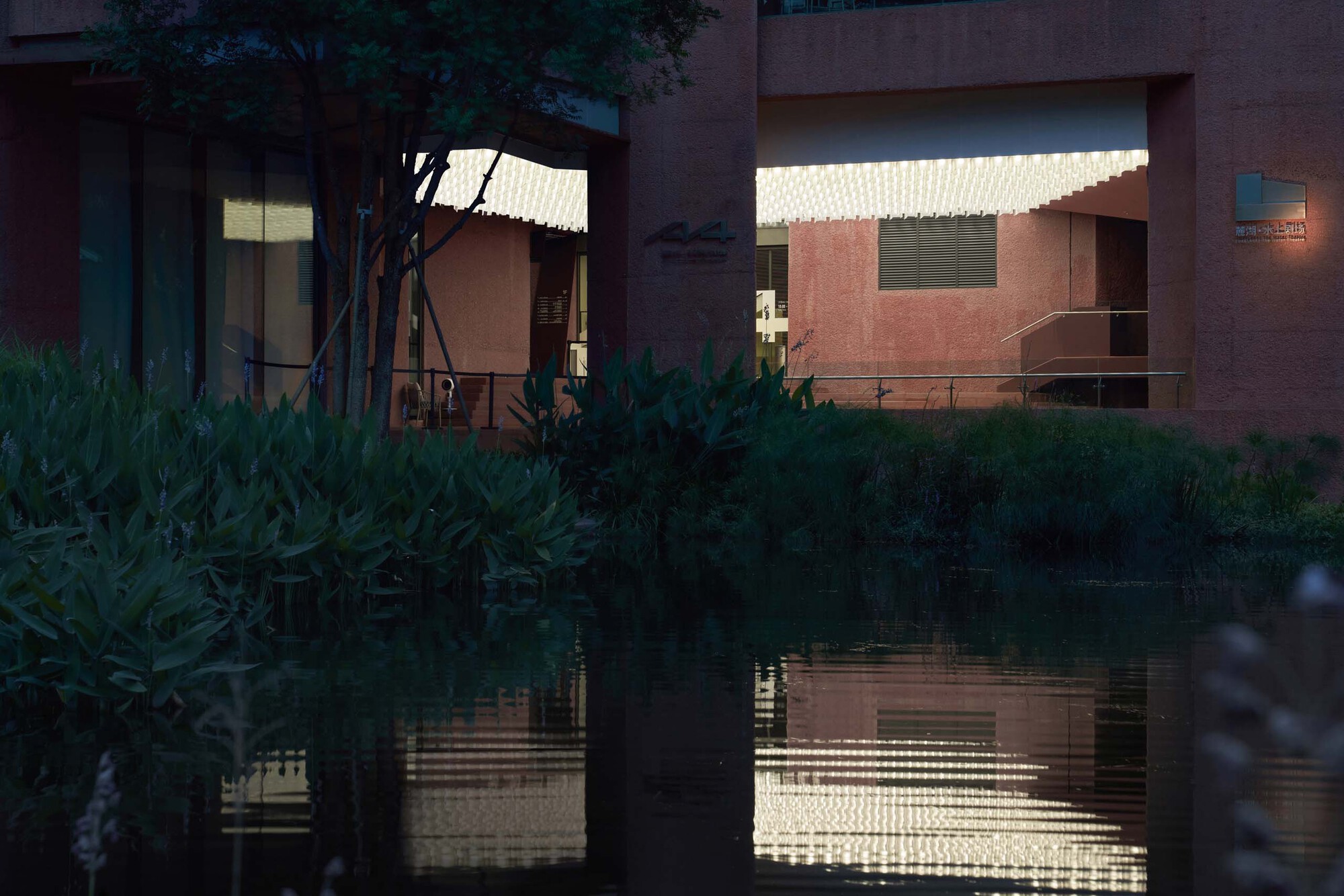 Evening view of illuminated interior volumes reflected in pond with lush foreground vegetation