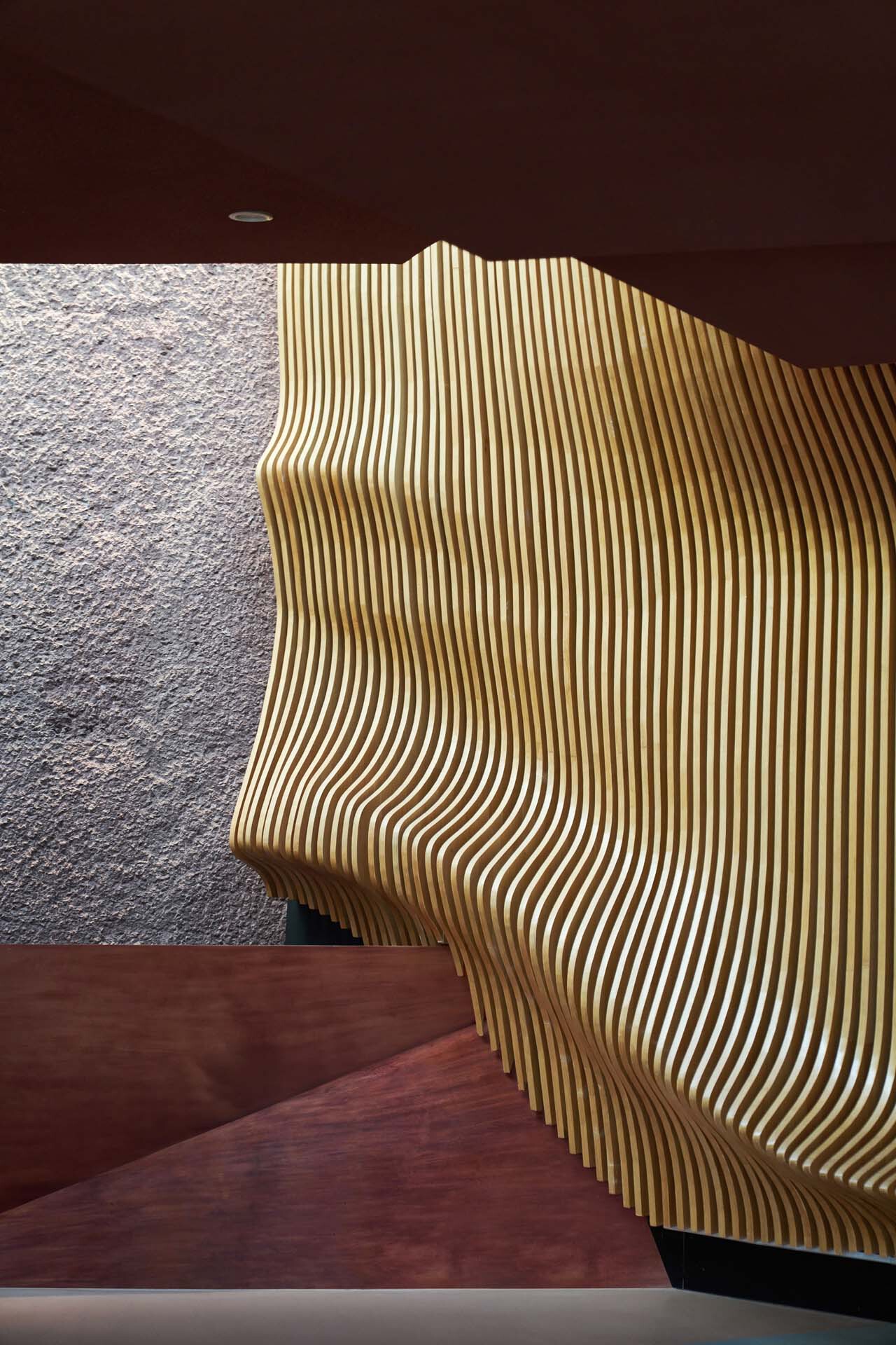 Curved wood slat wall meeting textured stone surface under corten ceiling in natural daylight