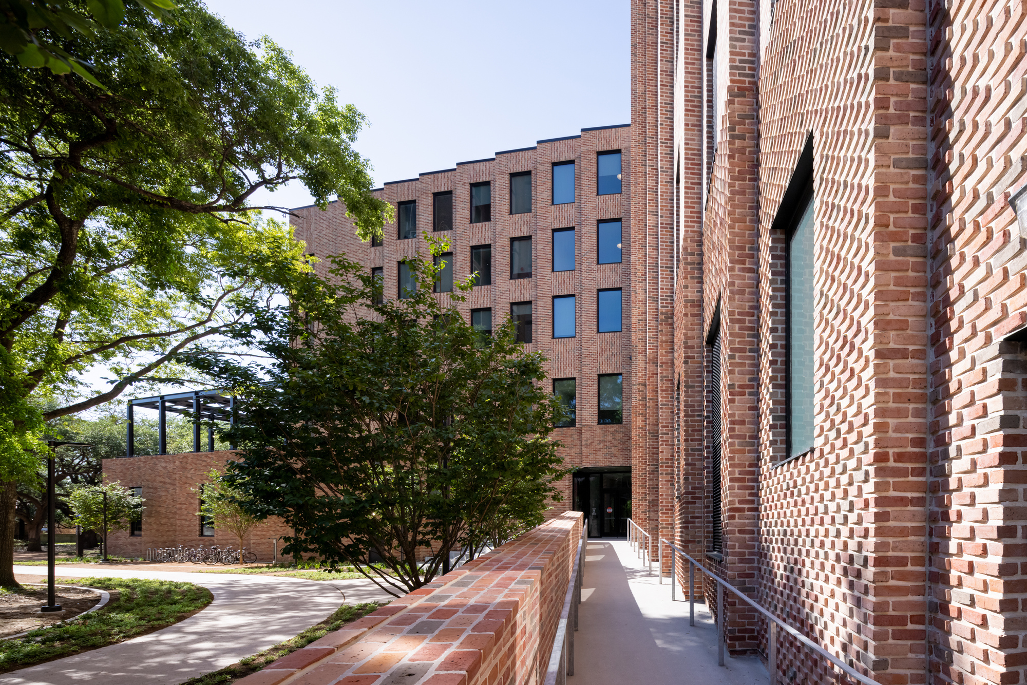 Patterned brick wall with vertical slits beside a shaded pathway and mature tree