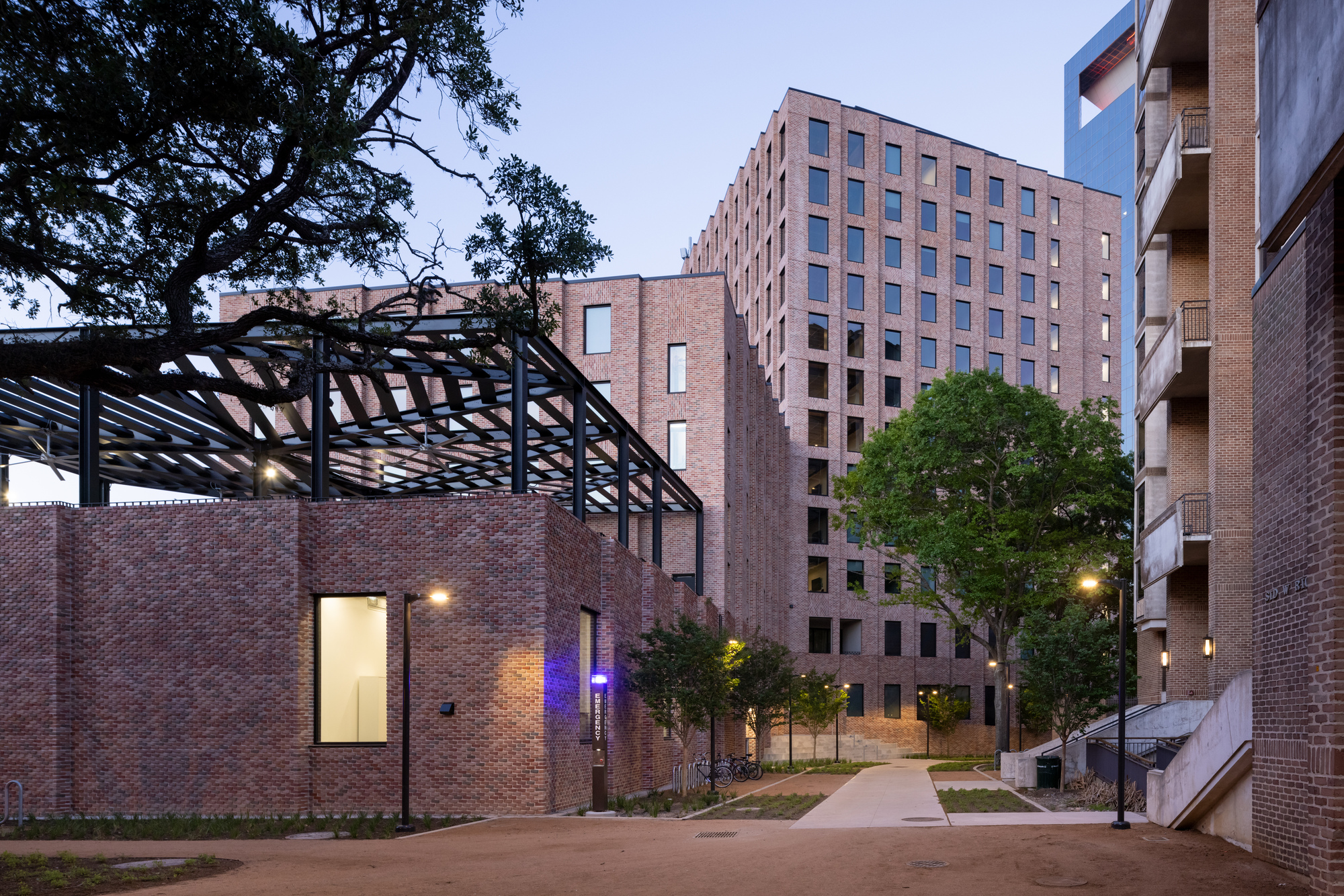 Courtyard passage at dusk with black steel pergola and illuminated brick walls between residential towers