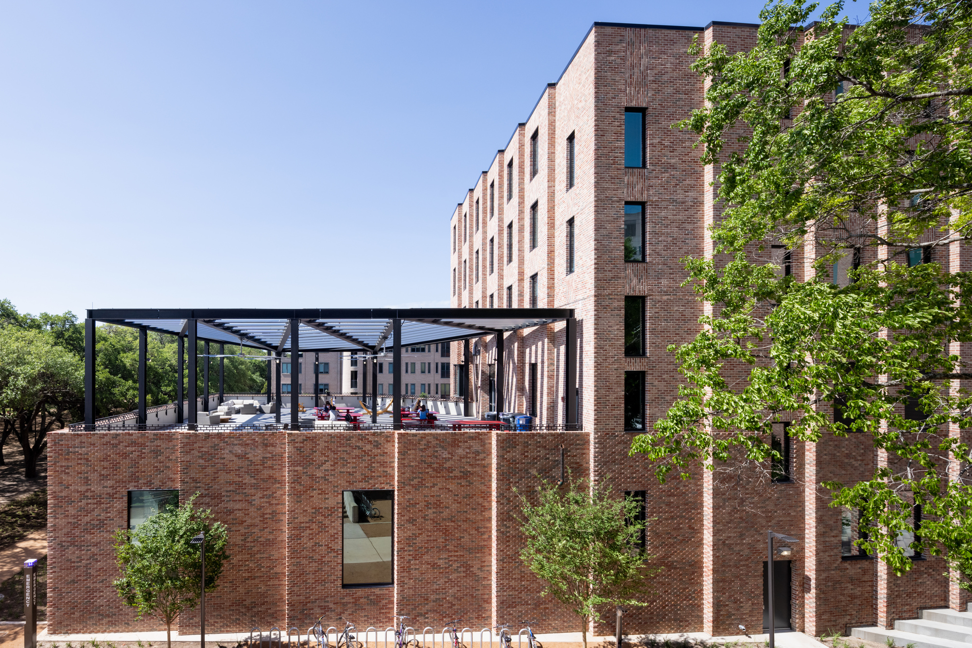 Single-story brick pavilion with steel pergola structure overlooking the courtyard with young trees in foreground