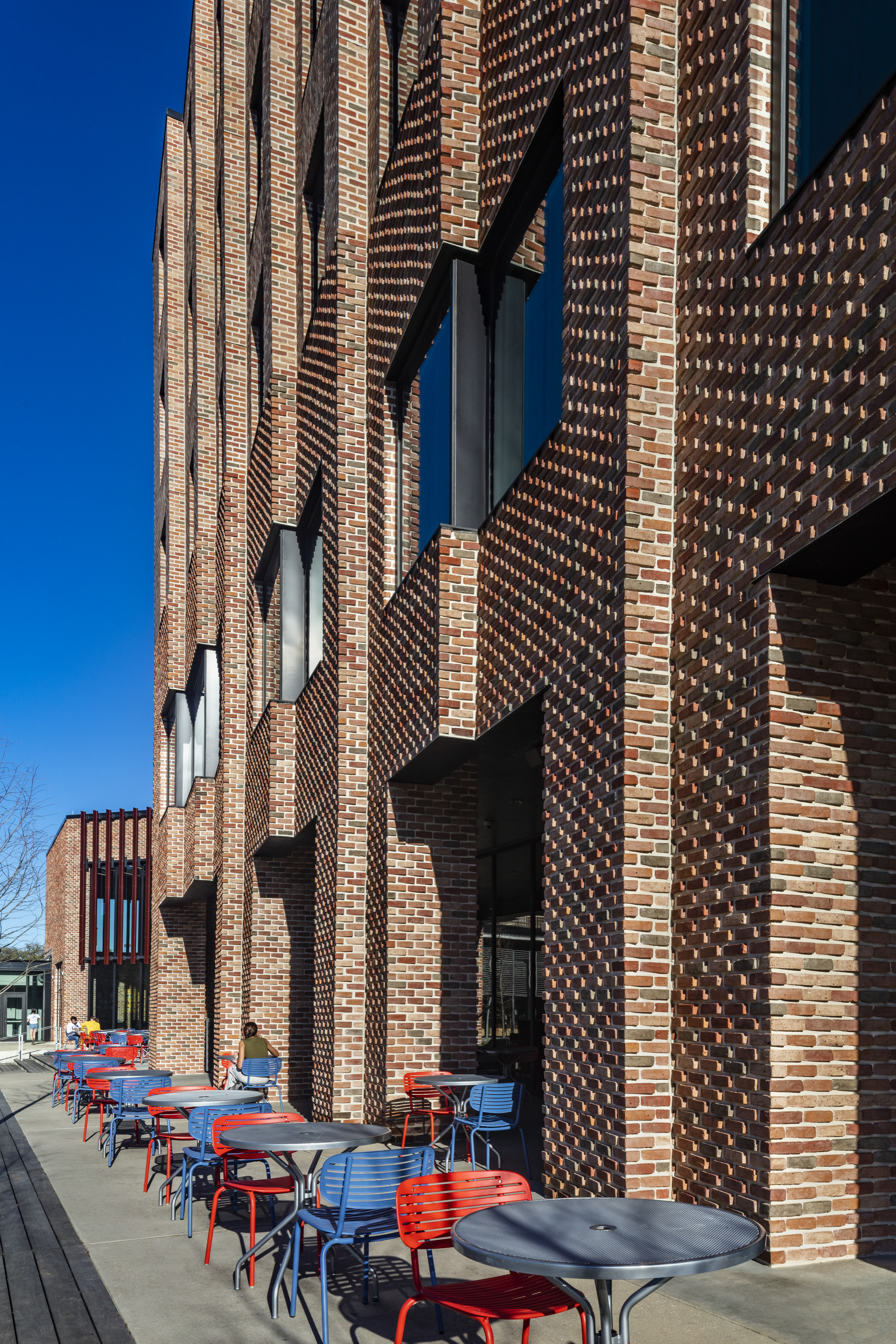 Textured brick facade with protruding and recessed patterns beside outdoor seating area with red and blue chairs