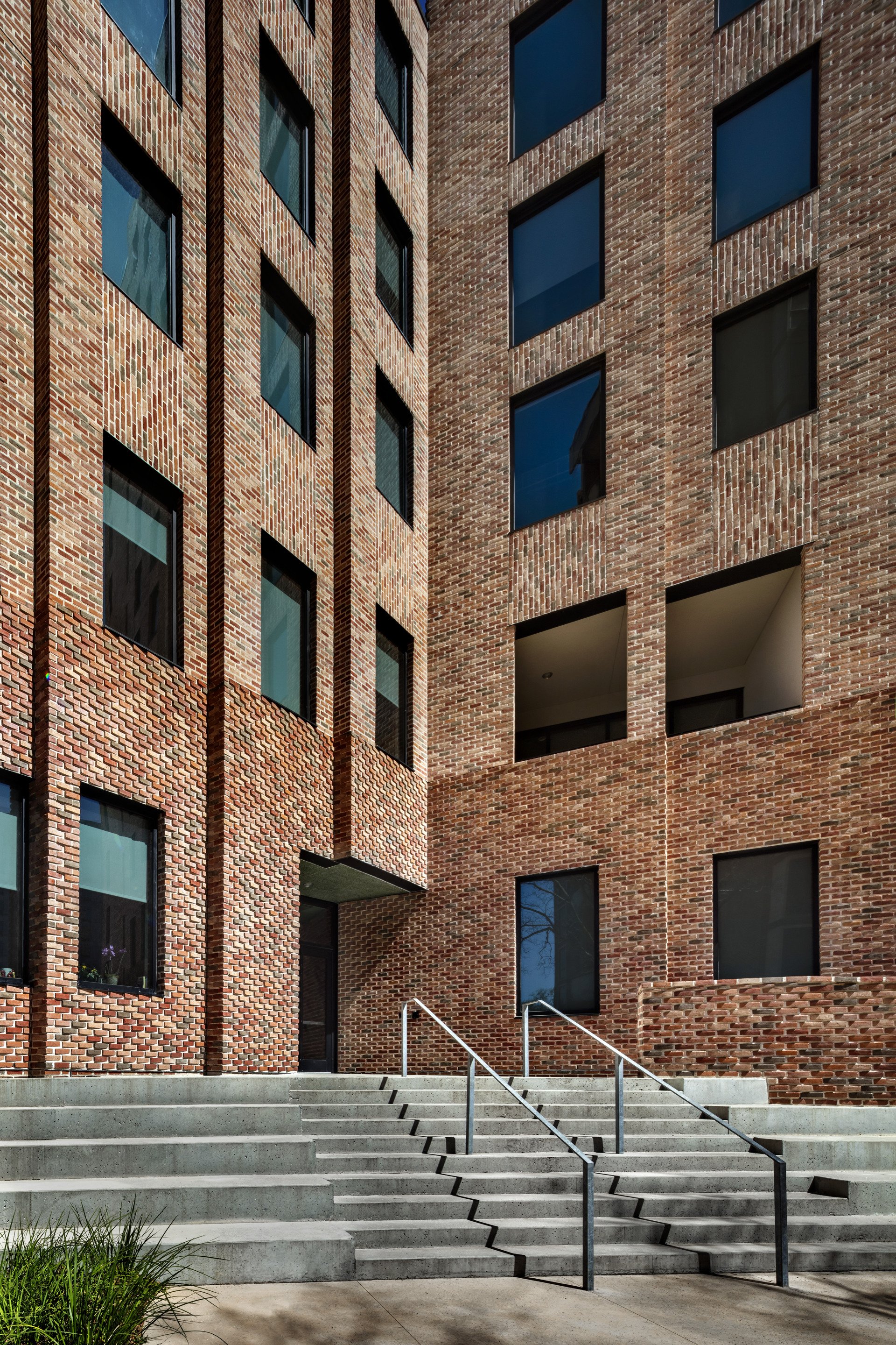 Concrete steps with metal handrails leading up to brick facade with punched window openings in afternoon sun