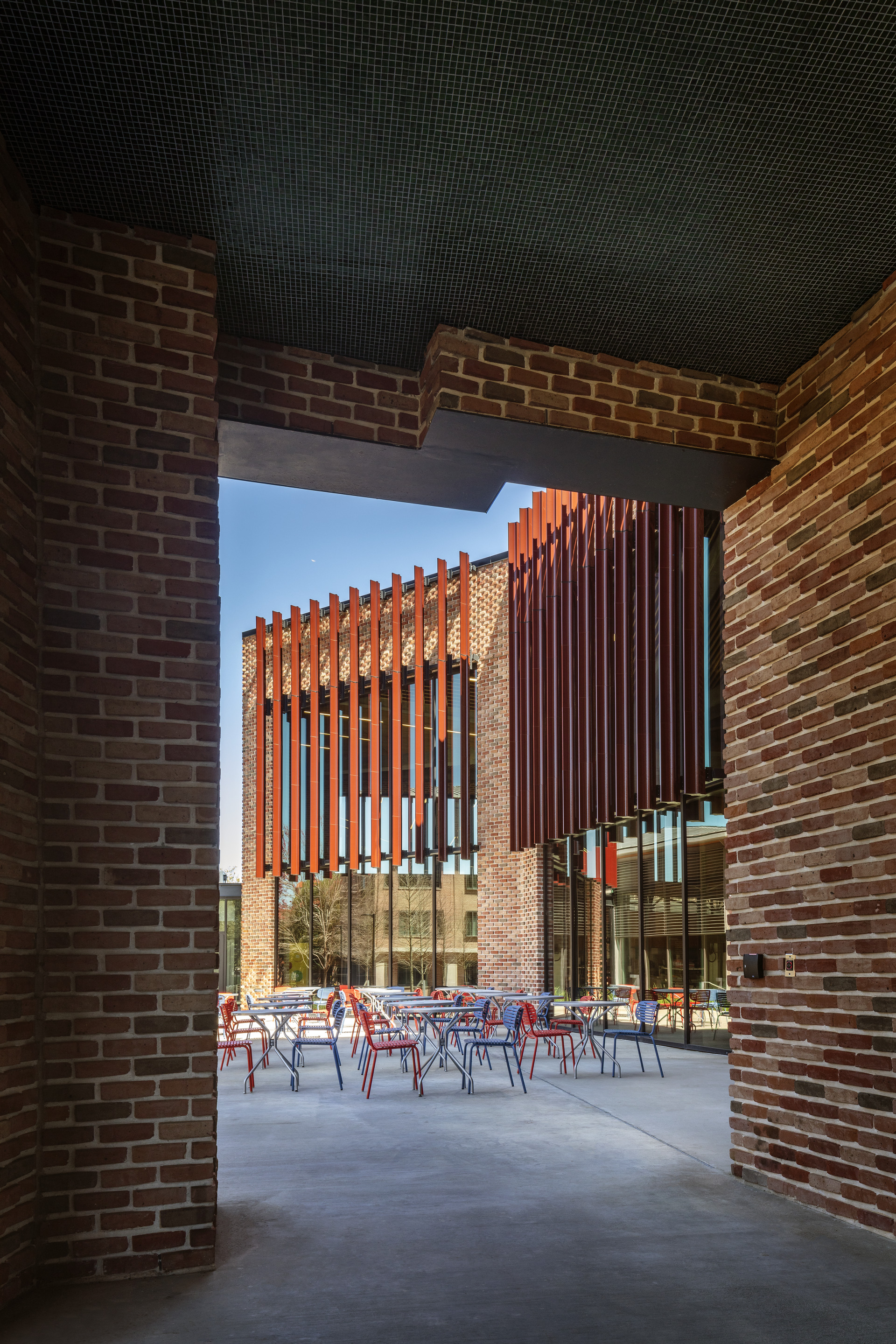 View through a brick-walled passageway into a courtyard with red chairs and vertical timber louvres beyond
