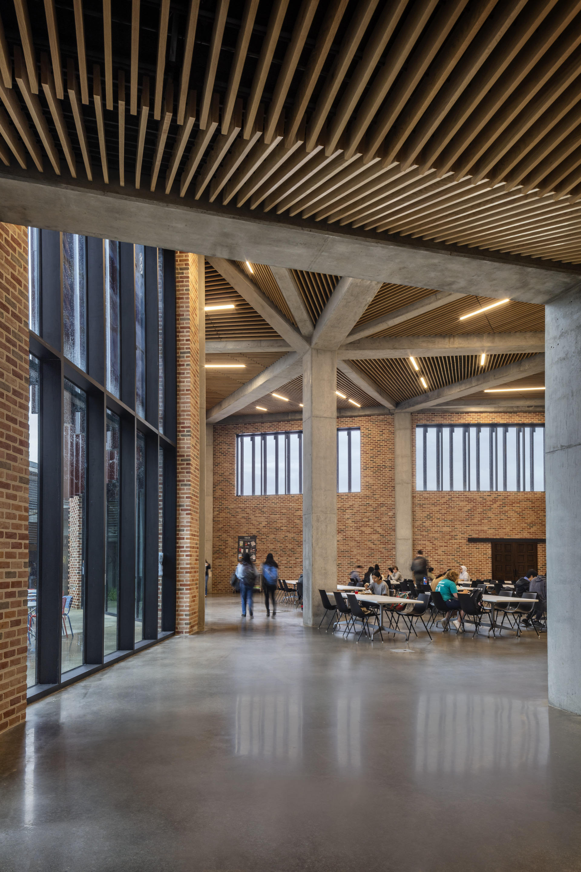 Concrete columns supporting a timber baffle ceiling above a polished floor with people seated in the background