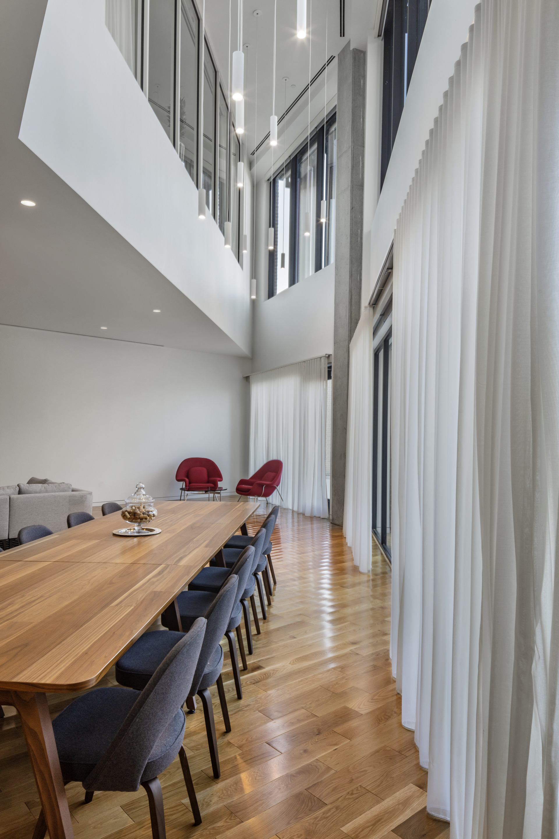 Double-height dining room with long timber table beside floor-to-ceiling sheer curtains and chevron wood flooring