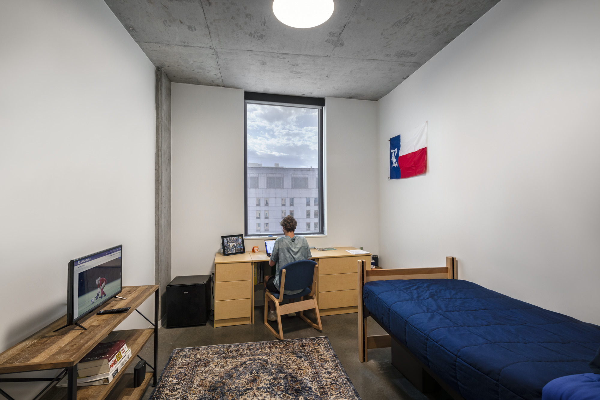 Dormitory room with exposed concrete ceiling, narrow window framing cloudy sky and neighboring buildings