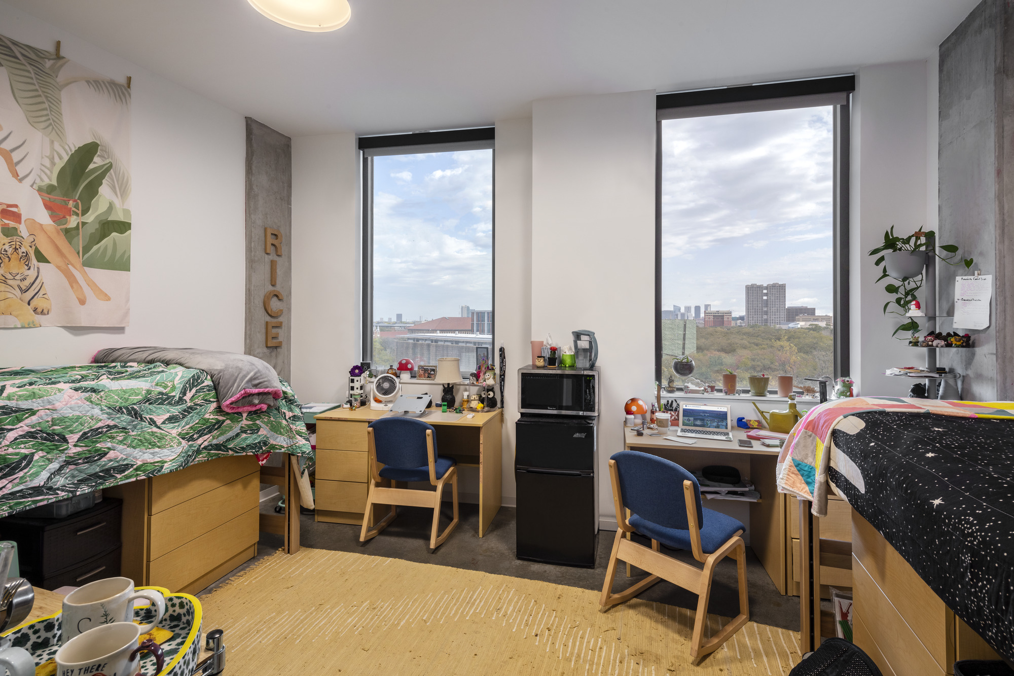 Shared dormitory room with corner windows overlooking the city skyline under partly cloudy skies