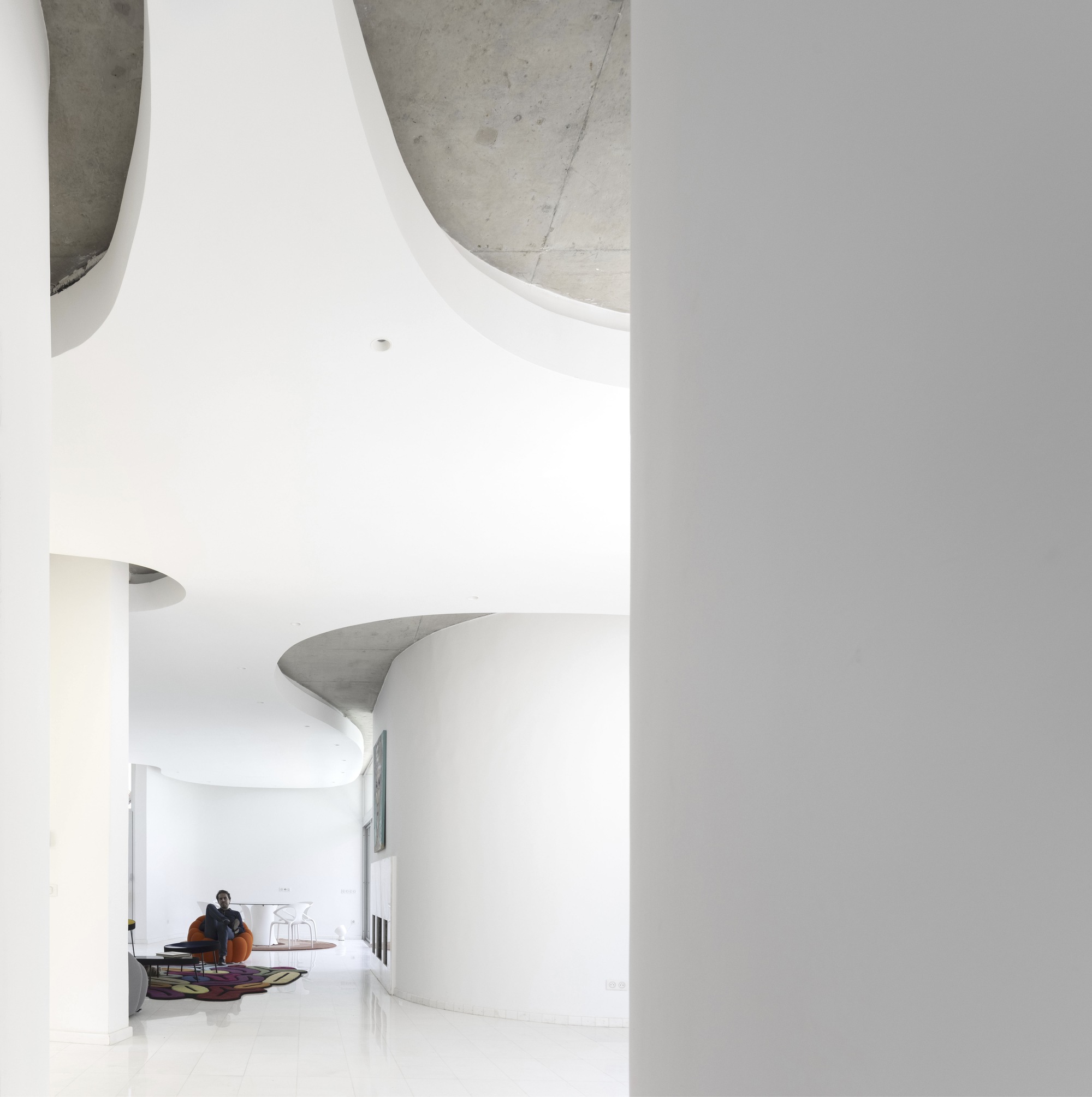 Curved white wall meeting a sculptural concrete ceiling element with a seated figure in the background