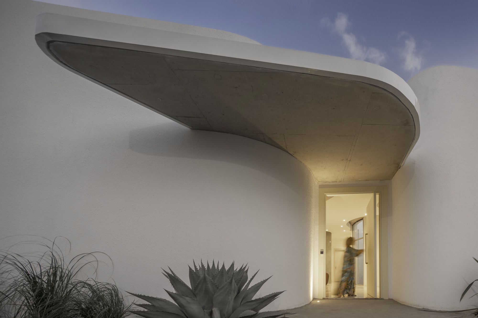 Illuminated entrance beneath a curved concrete canopy with agave and grasses at dusk