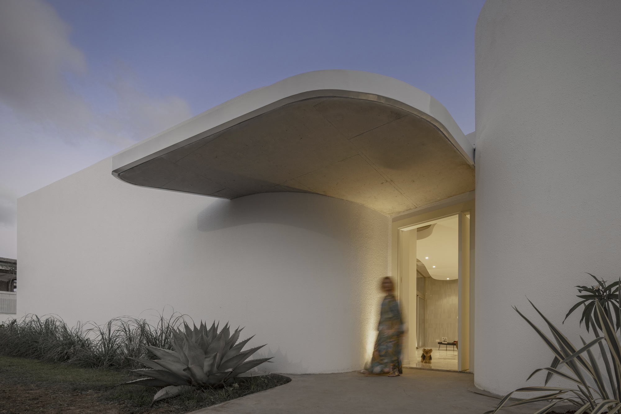 White stucco entry wall with sweeping concrete soffit and figure passing through at twilight