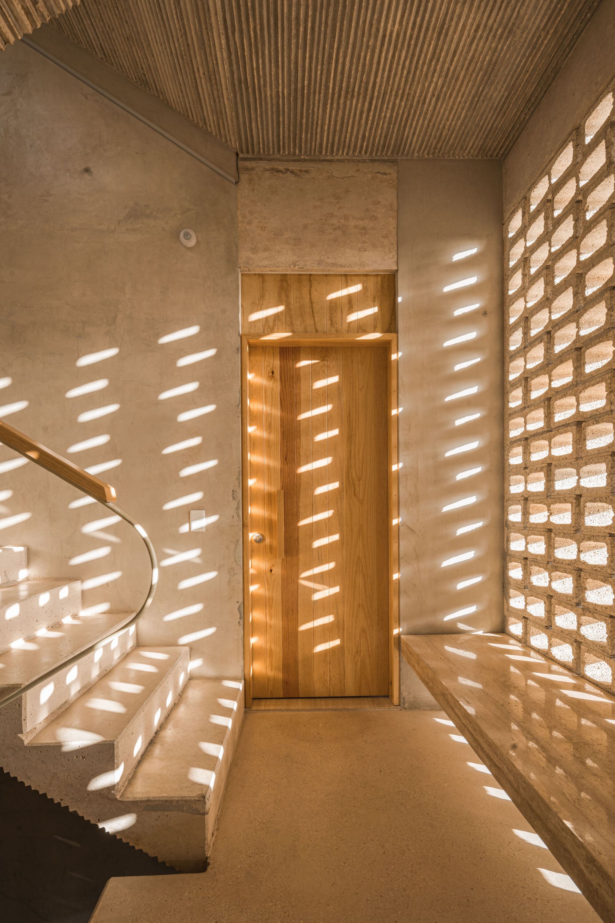 Entry foyer with perforated brick screen filtering dappled sunlight across concrete stairs and timber slatted ceiling