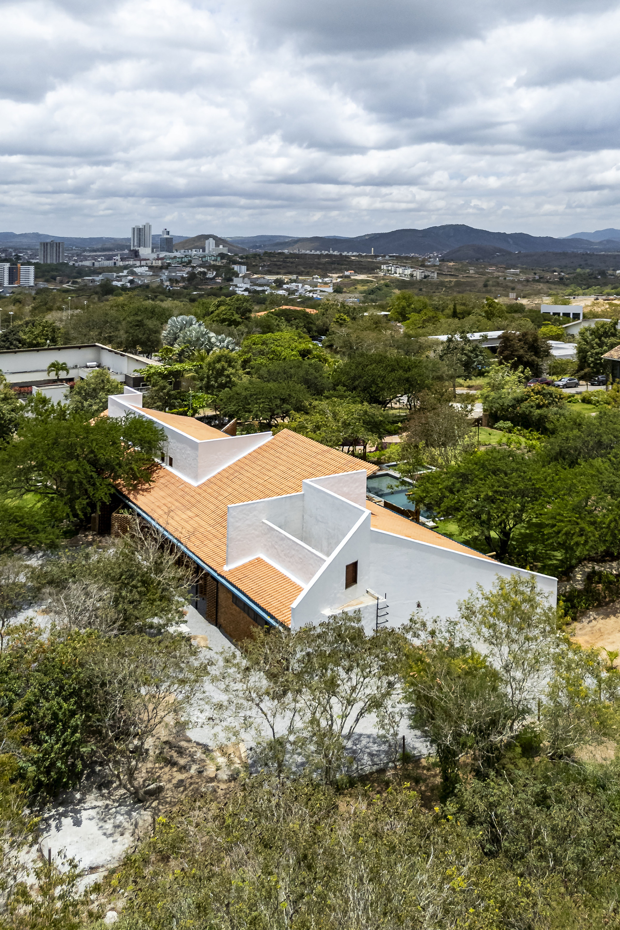 Angular orange tile roof with white parapets set among mature trees under cloudy skies