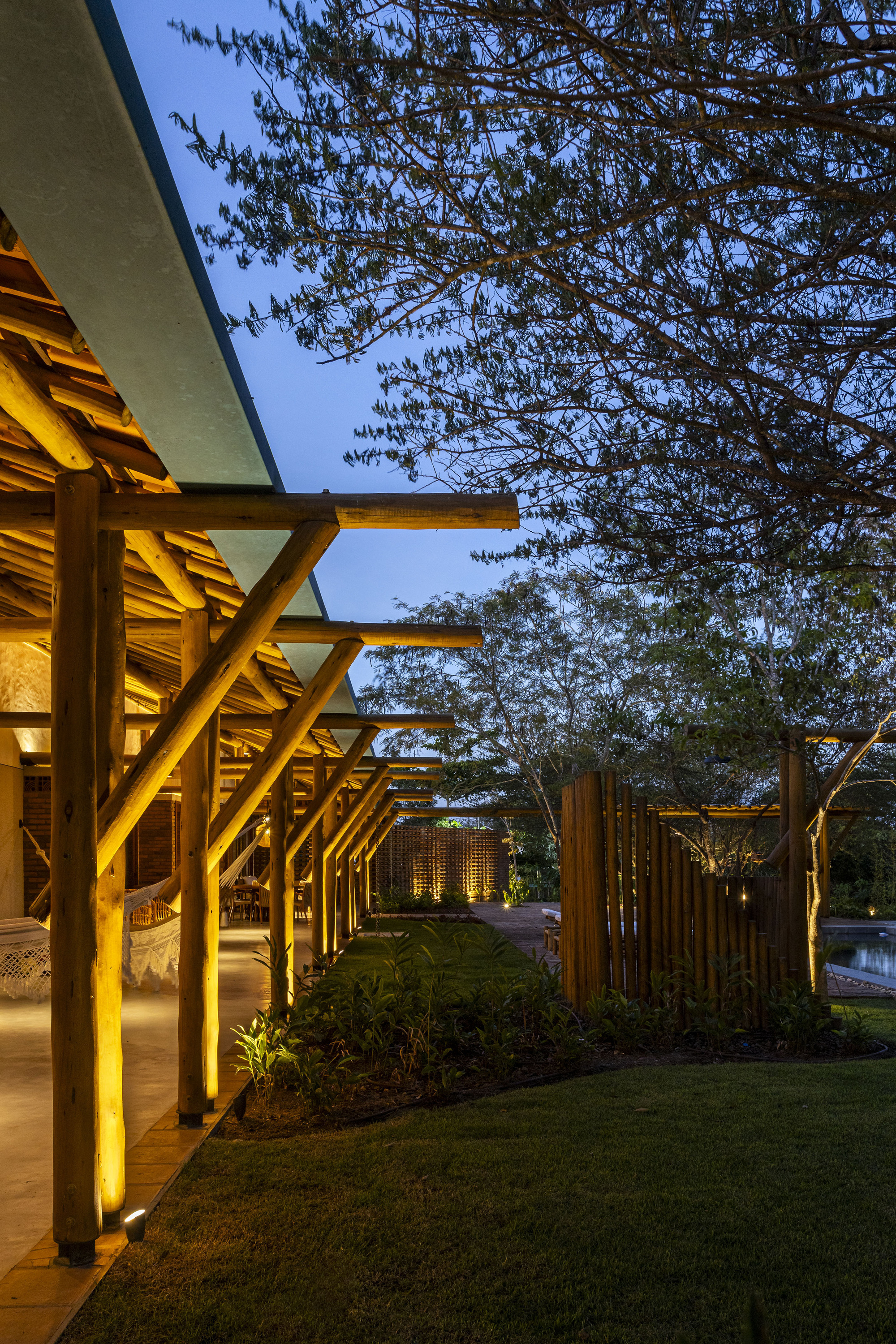 Illuminated timber colonnade with exposed beams supporting a flat roof beside lawn and mature trees at dusk