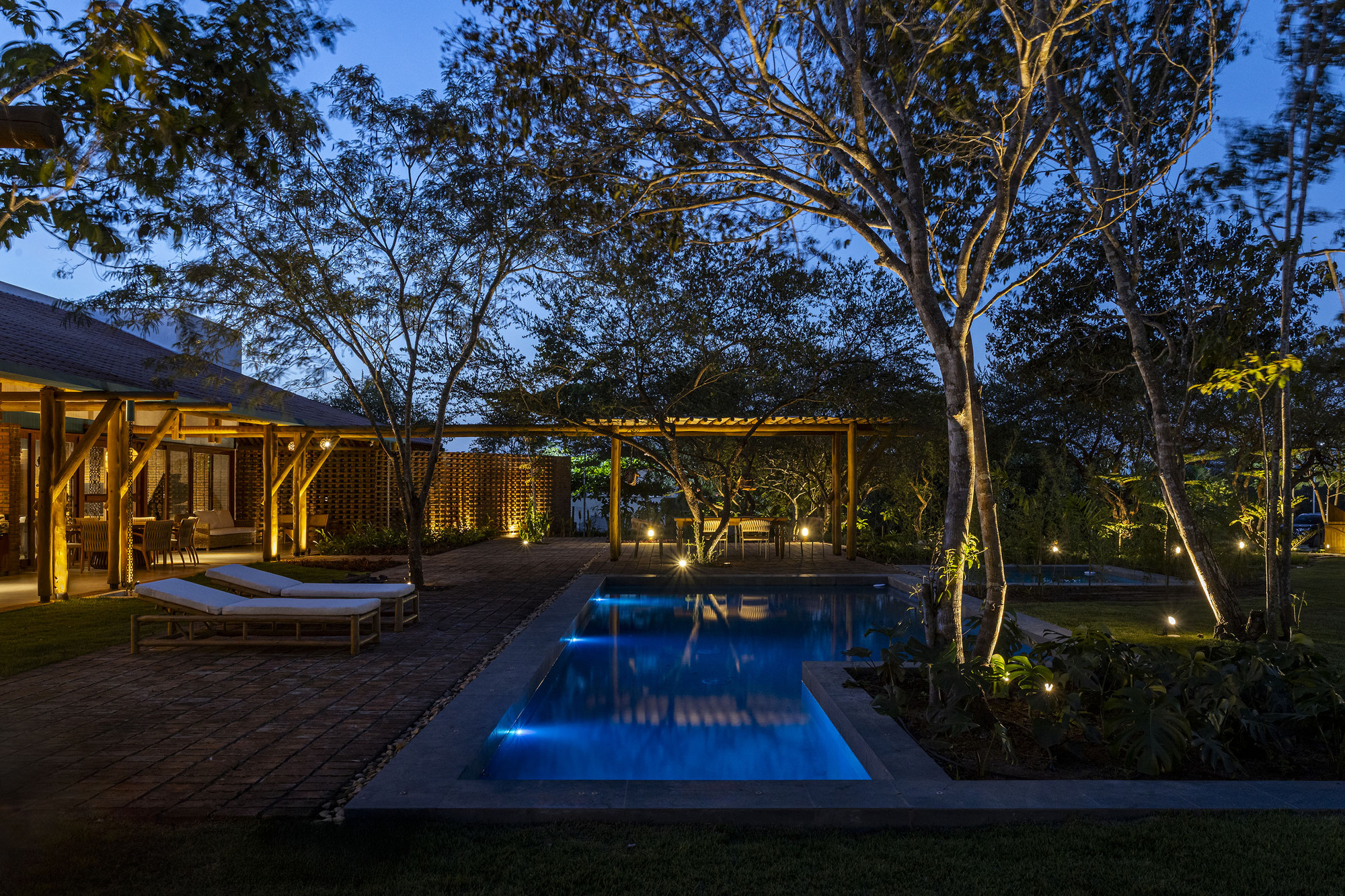 Pool terrace with timber deck and loungers framed by mature trees and uplighting at blue hour
