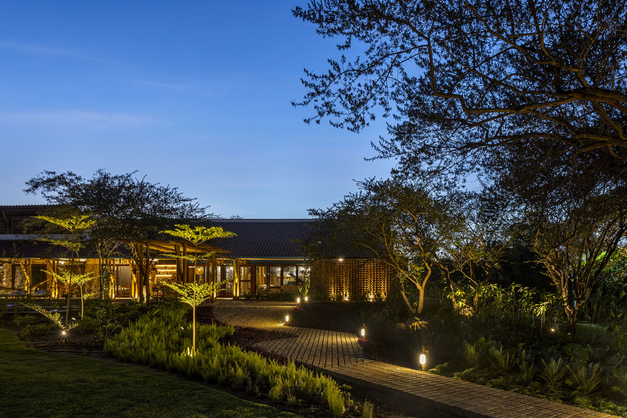Low-slung pavilion with corrugated roof and timber screen walls along brick paved path through landscaped garden at dusk