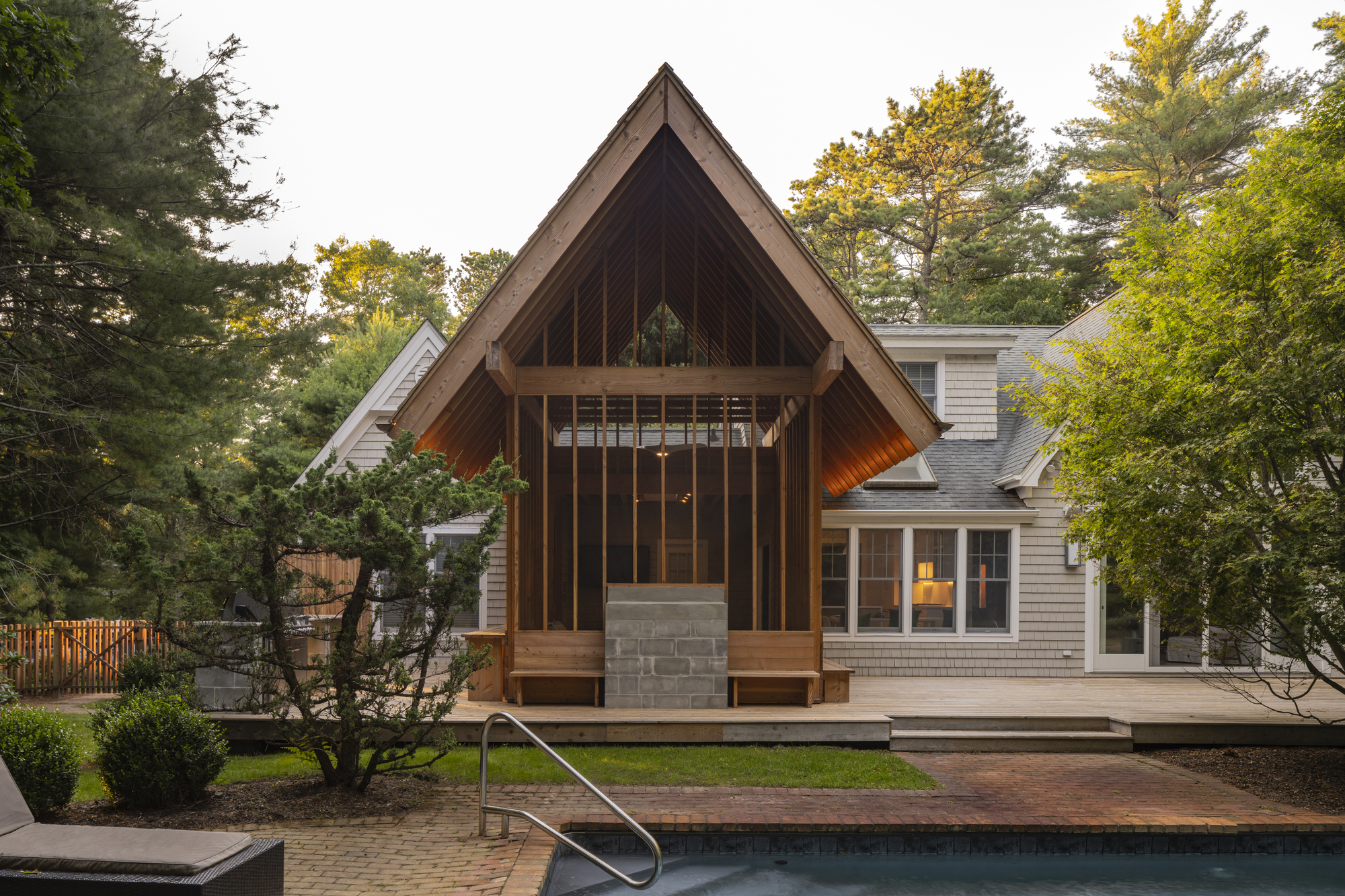 Timber pavilion with steep gabled roof and vertical slat facade beside a swimming pool at dusk