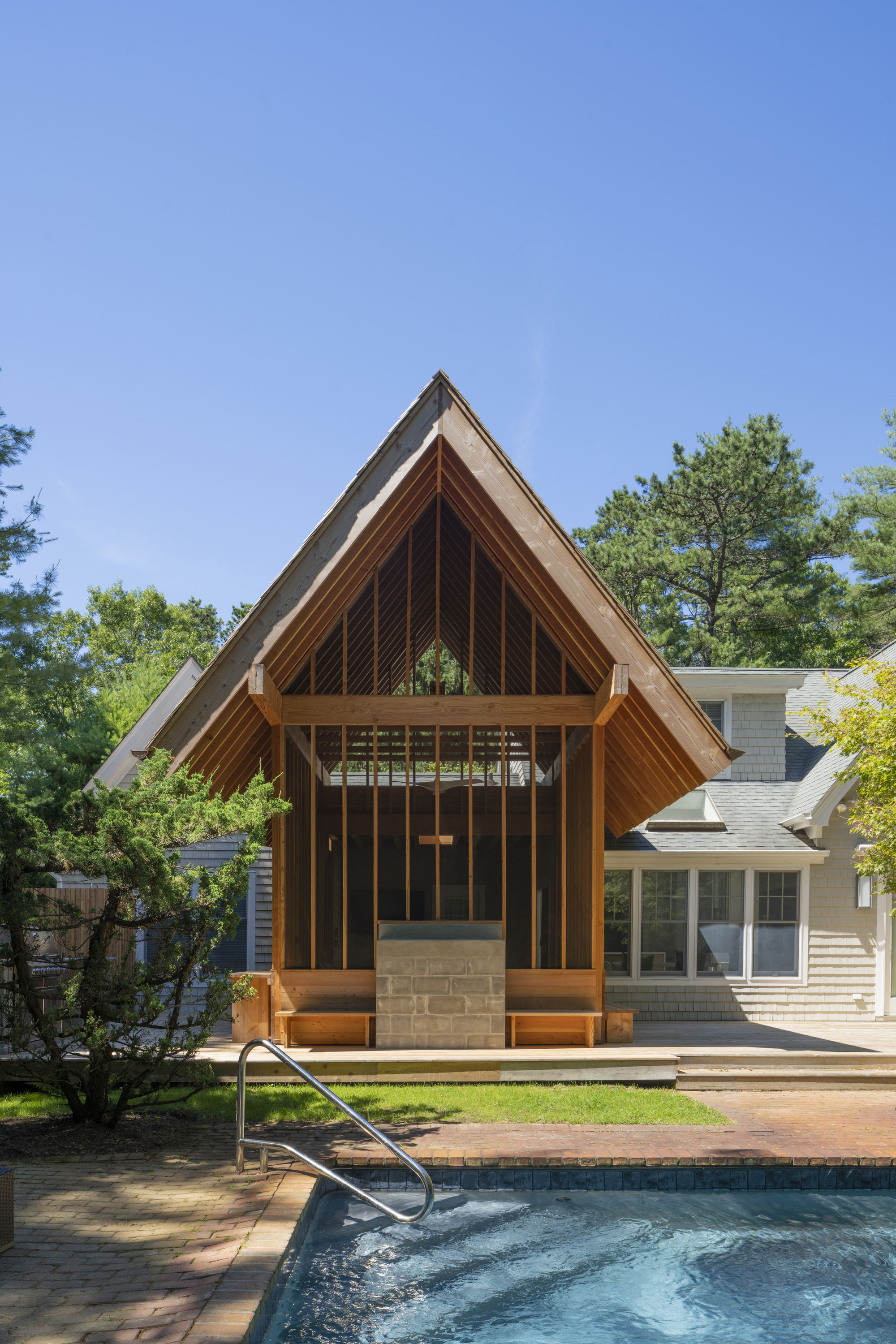 Timber-framed pavilion with steep gable and vertical slats overlooking a swimming pool on a summer day