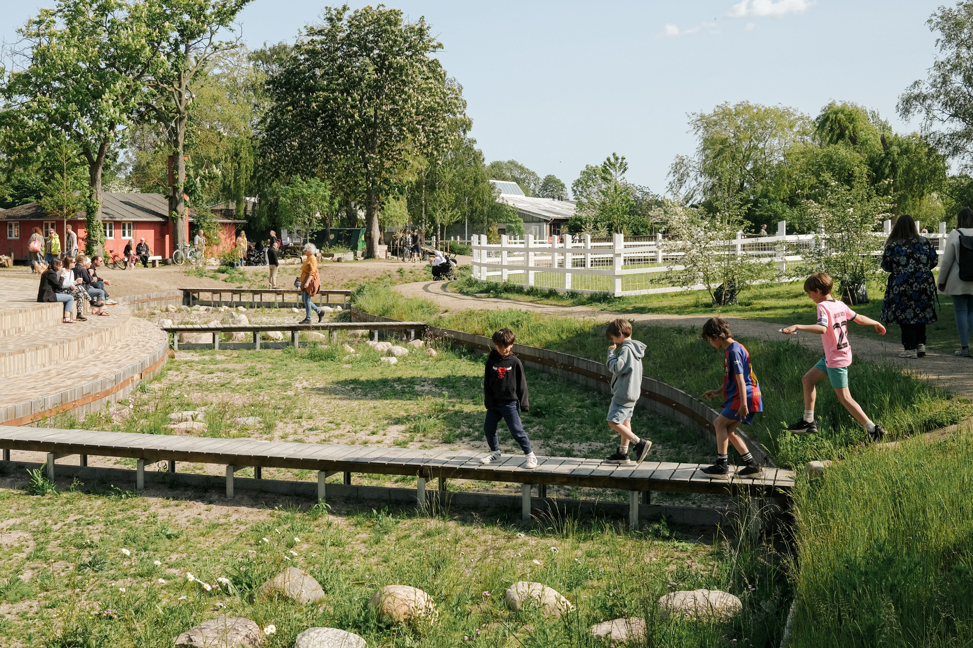 Children crossing a low wooden footbridge over a planted bioswale bordered by timber benches