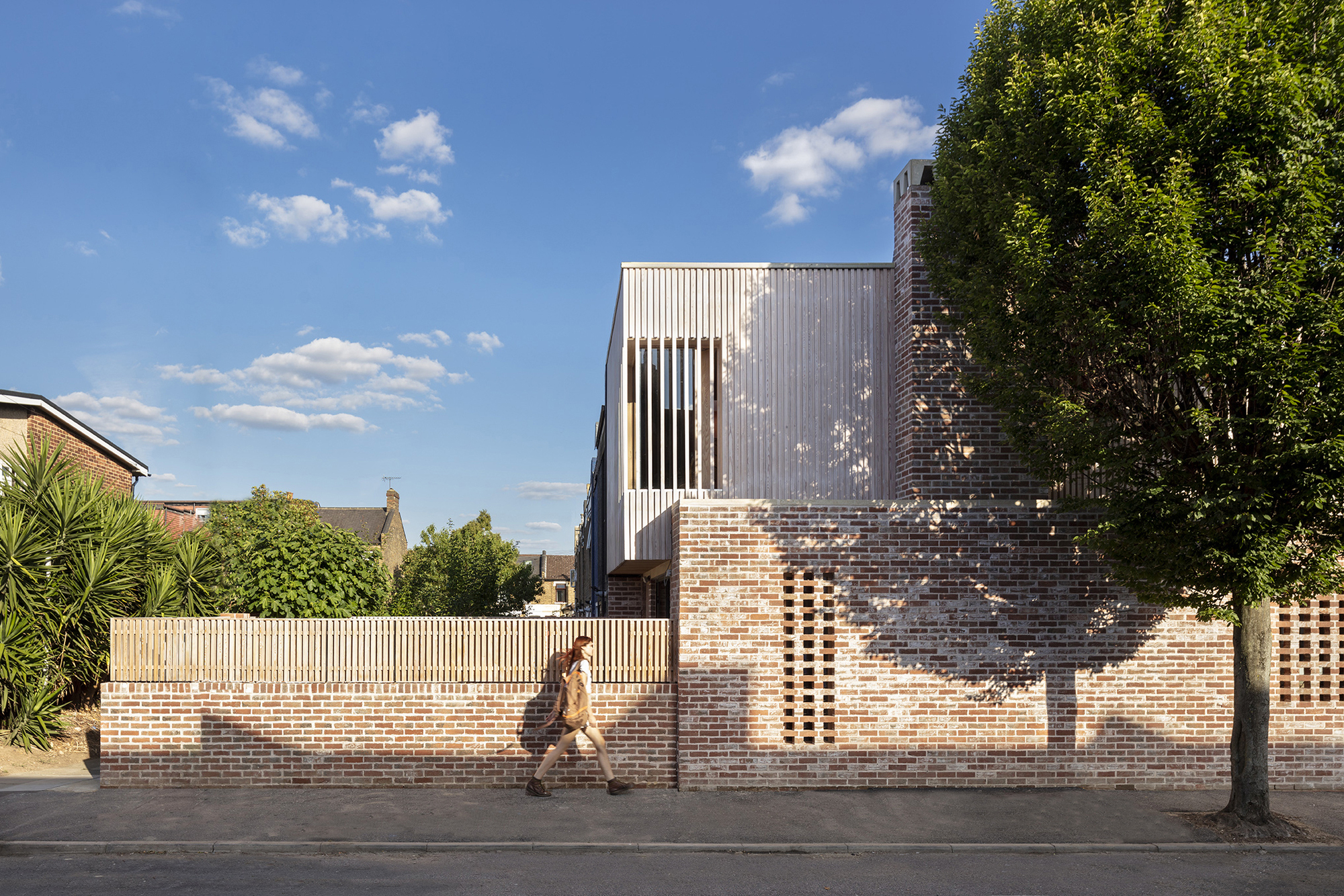 Street view of perforated brick wall with timber fence and corrugated metal upper volume as a person walks past