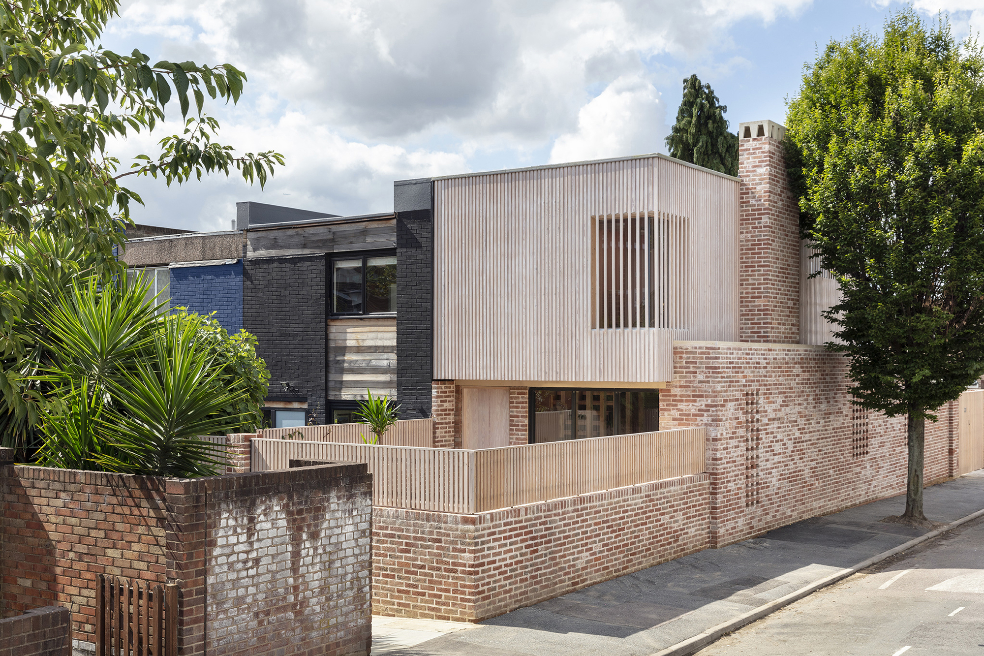 Facade showing corrugated metal upper story above perforated brick walls with timber fencing and street trees