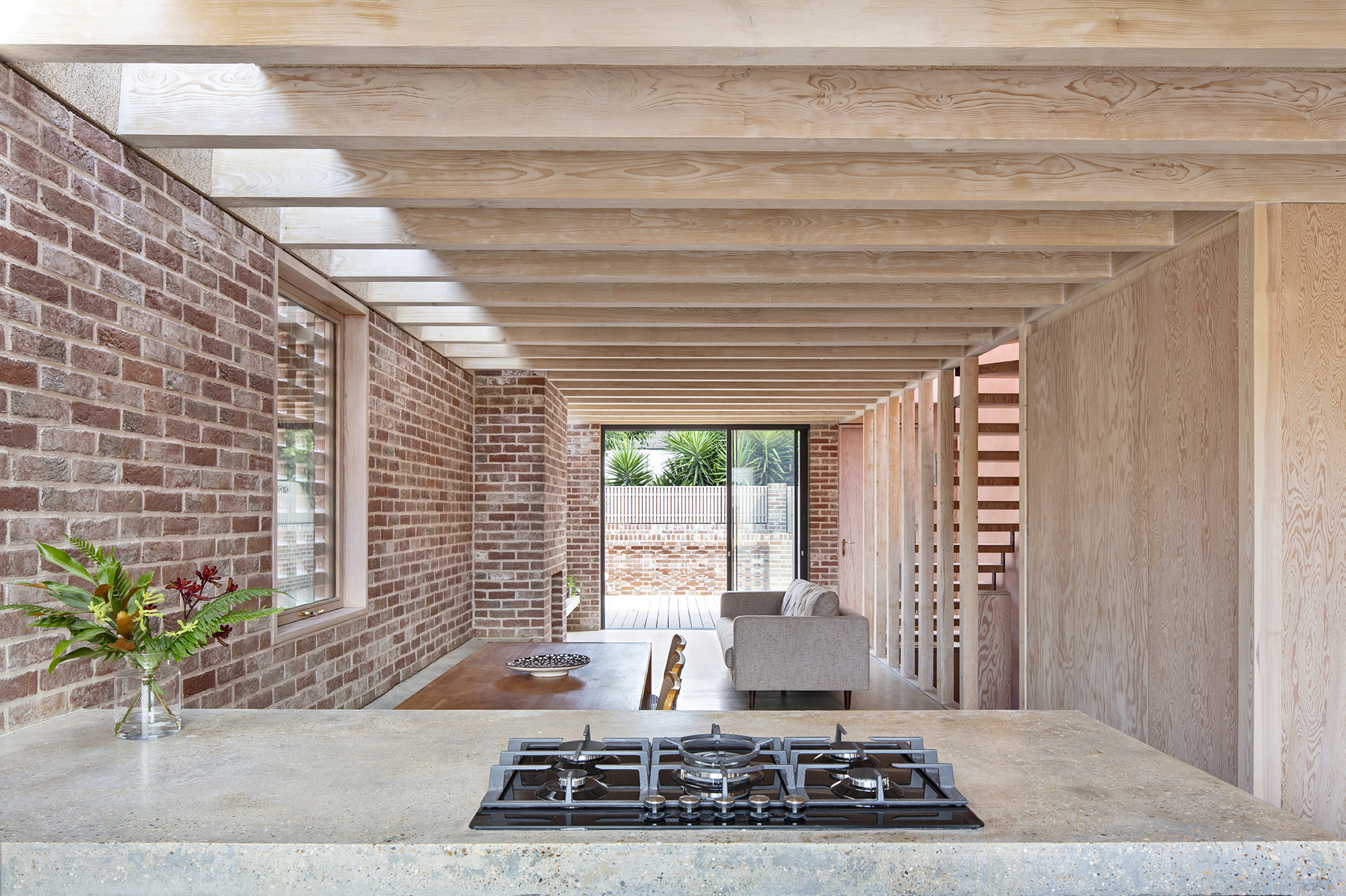 Open kitchen with exposed timber joists above concrete counter and gas cooktop facing brick walls