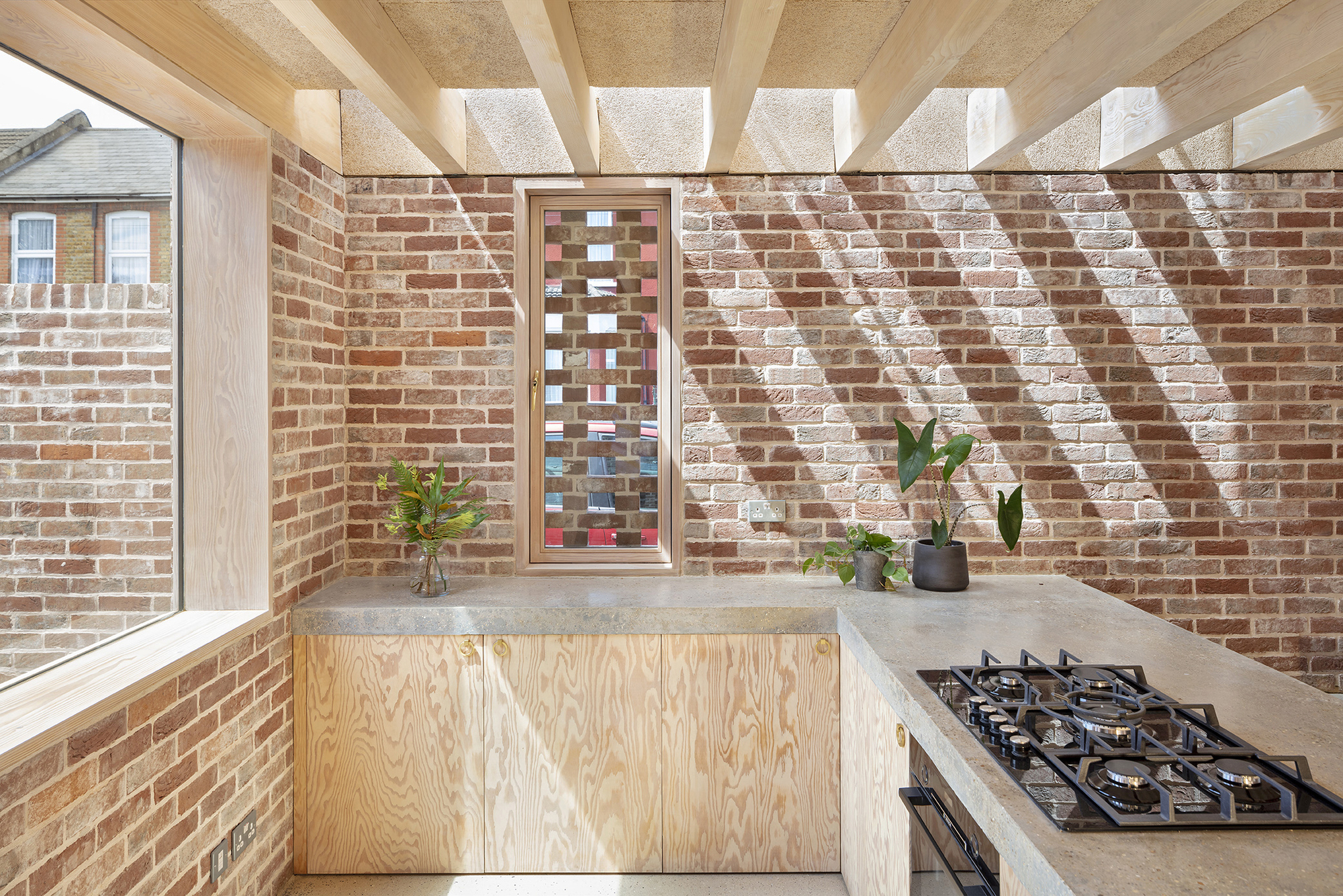 Kitchen corner with plywood cabinetry and slotted vertical window casting diagonal shadows across brick wall