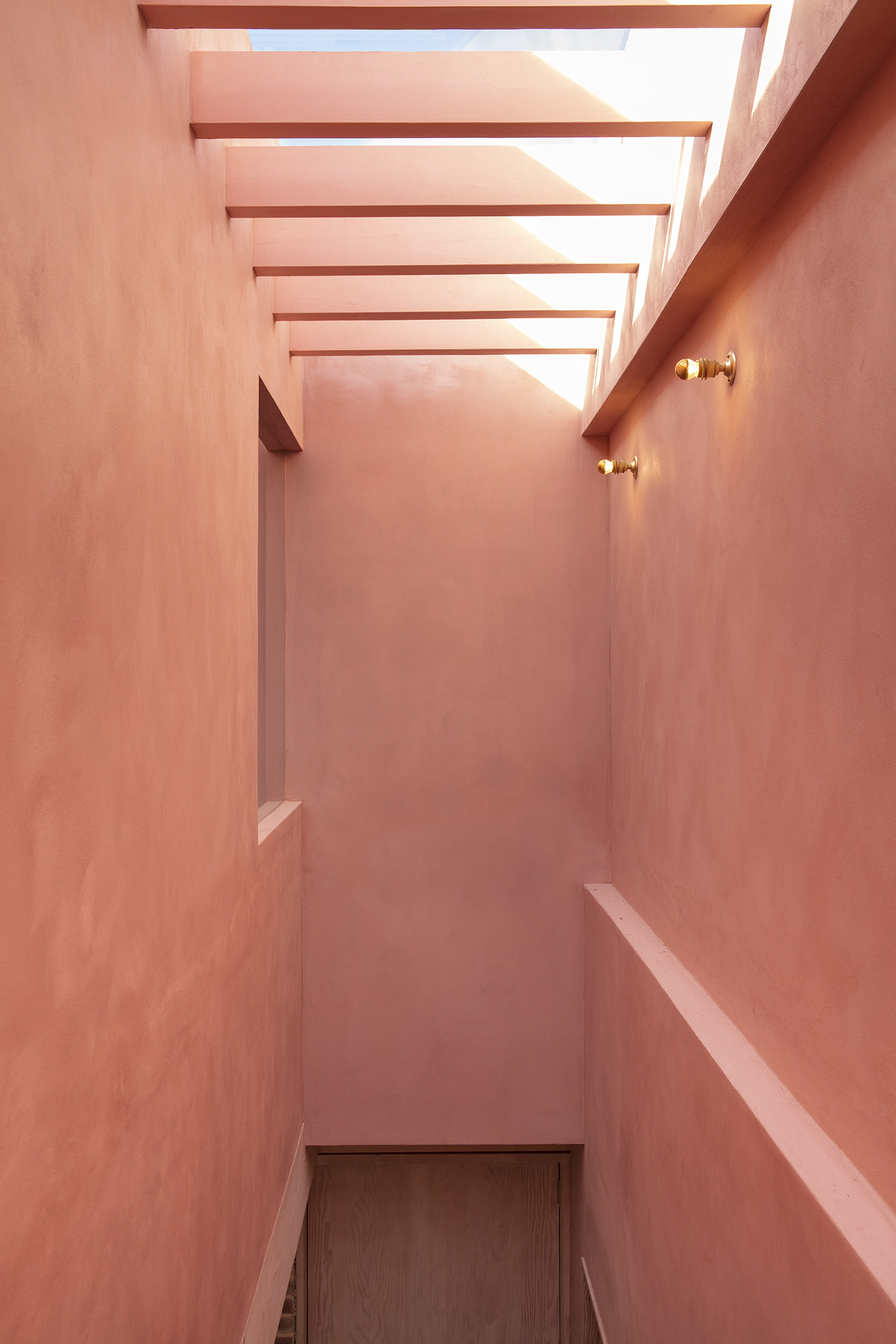 Narrow pink plastered stairwell with horizontal timber ceiling slats and brass wall sconces