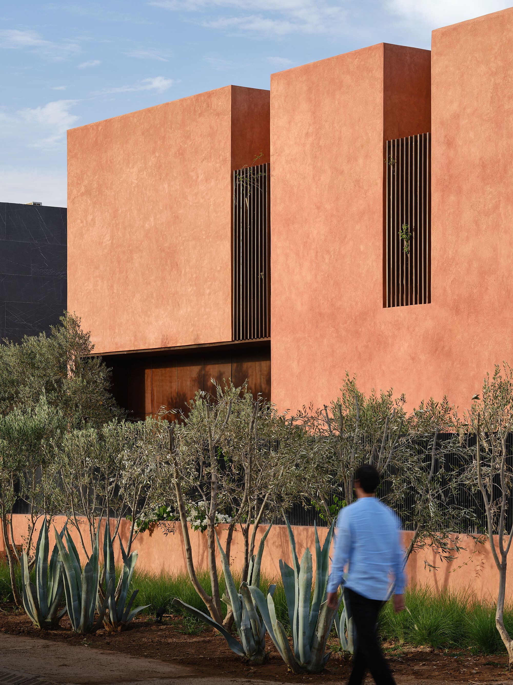 Corner of terracotta facade with vertical timber screens above olive trees and a passing pedestrian