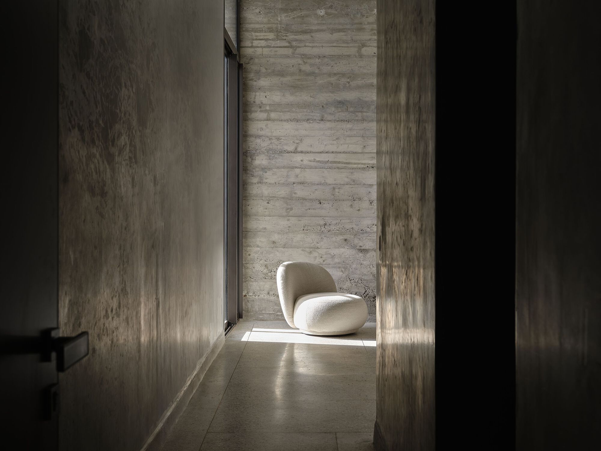 Narrow corridor with board-formed concrete walls and a single chair in sunlight