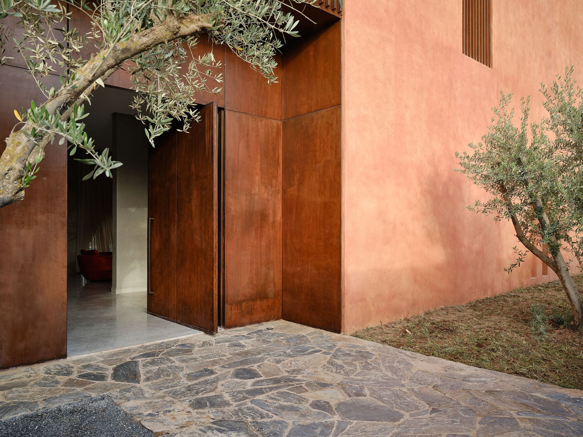 Courtyard entry with weathered steel pivot door framed by pink stucco walls and olive trees