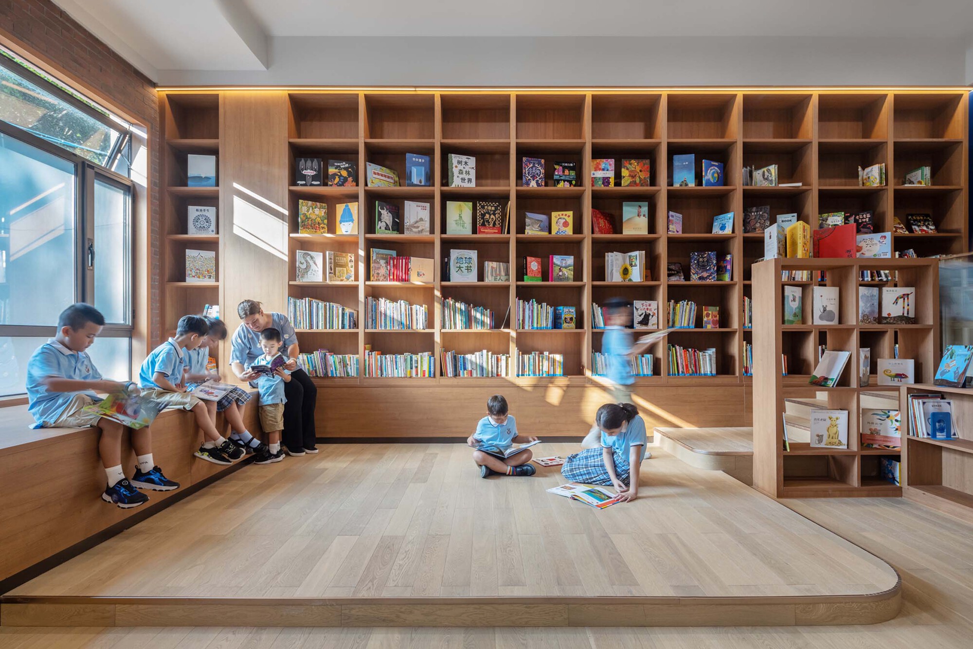Library with floor-to-ceiling timber bookshelves and stepped seating where children sit reading in morning light