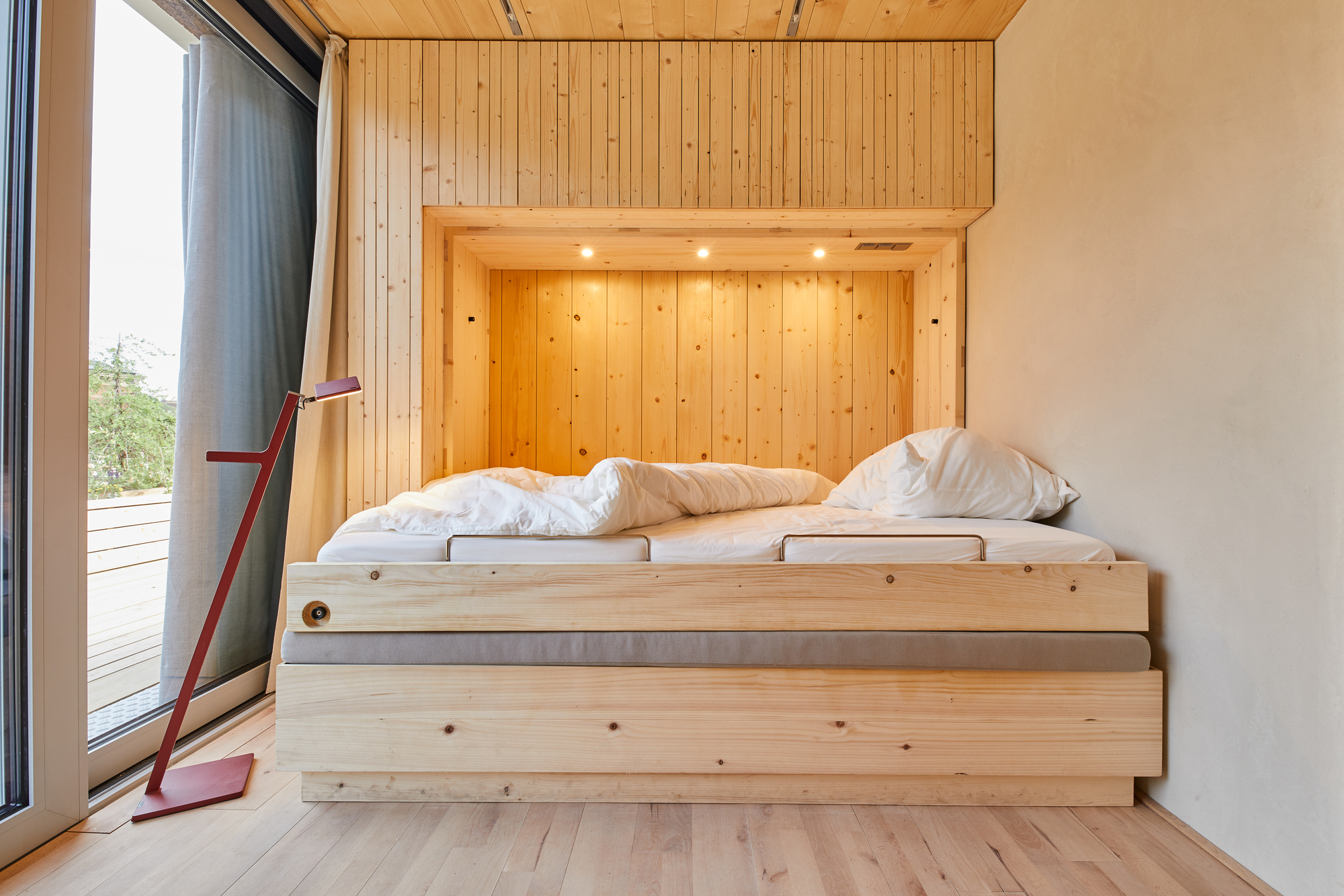 Bed alcove with white linens and plywood frame beside a glazed door