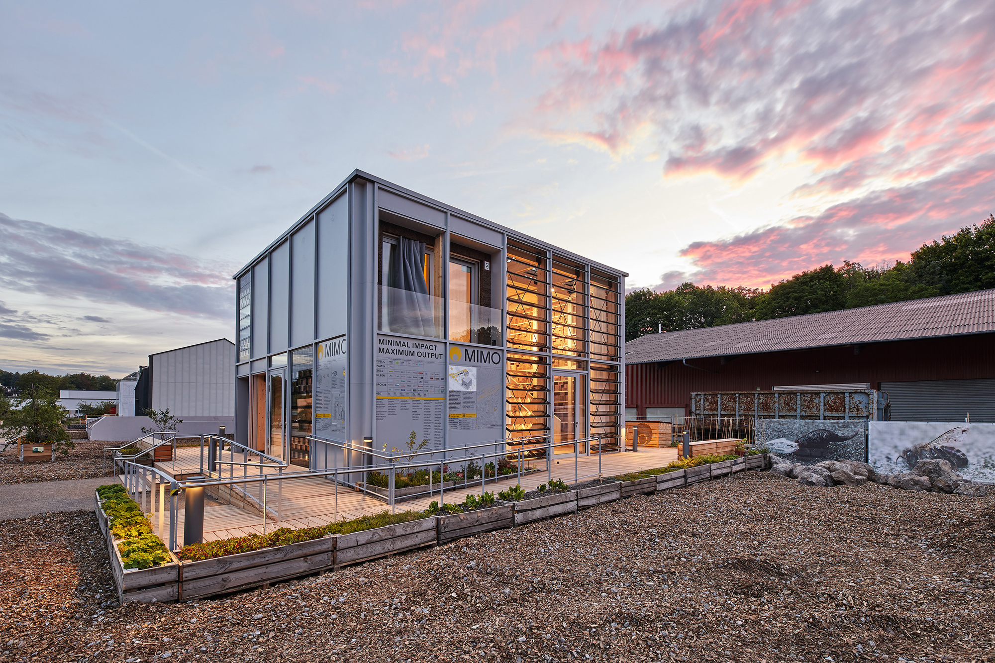 Two-storey corrugated metal structure with timber screen facade and planted beds under a pink sunset sky