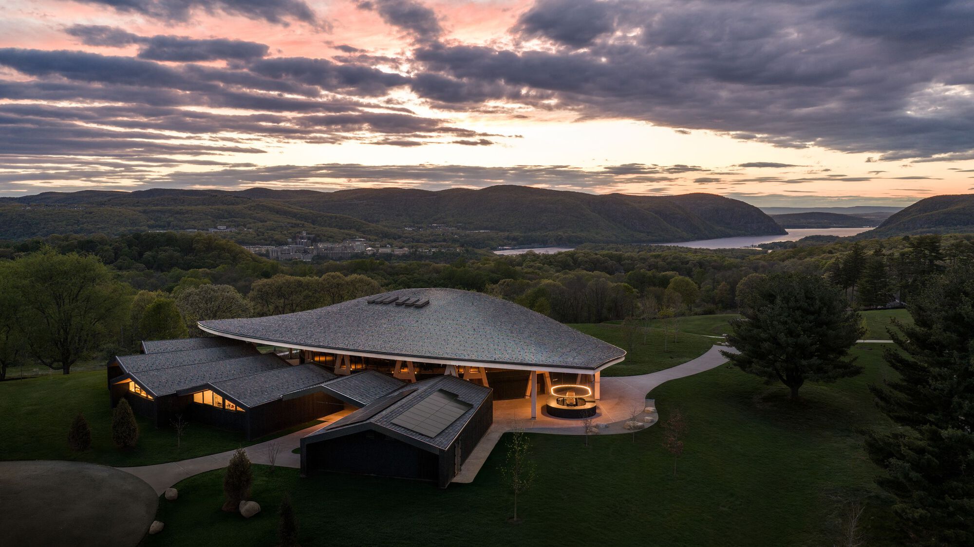 Aerial view of the curved roof volumes set in a lawn overlooking a lake at dusk