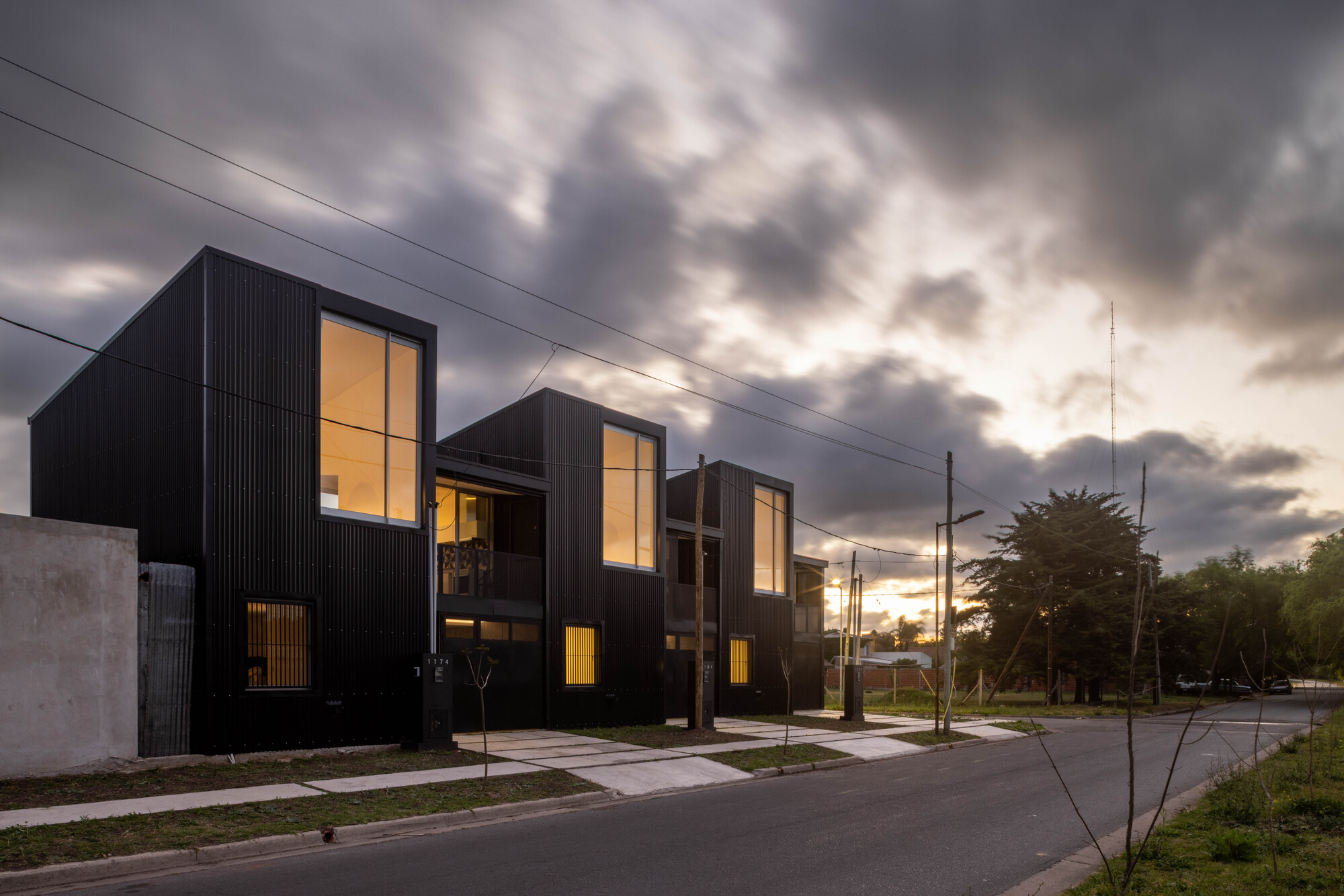 Facade of black corrugated townhouses with glowing interiors visible through tall windows at twilight