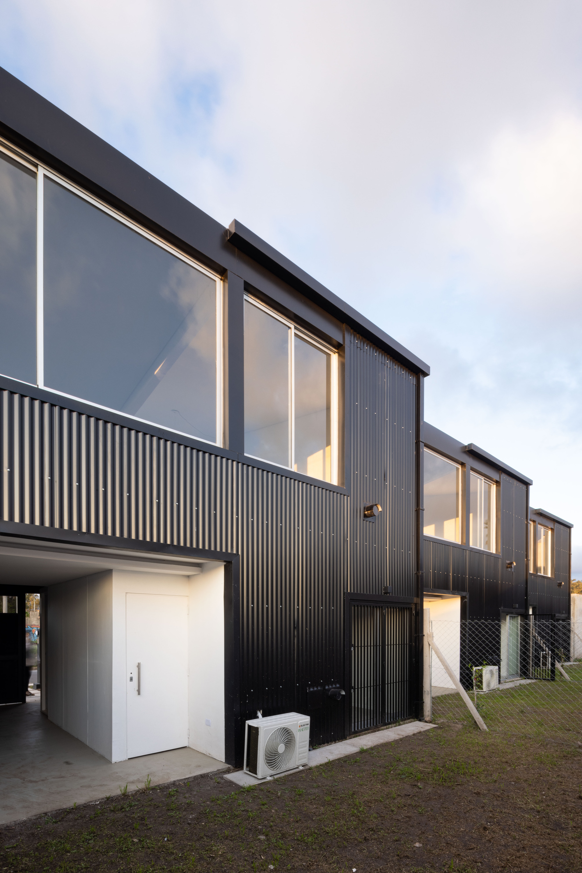 Row of black corrugated metal facades with upper glazing and ground-level carports under overcast sky