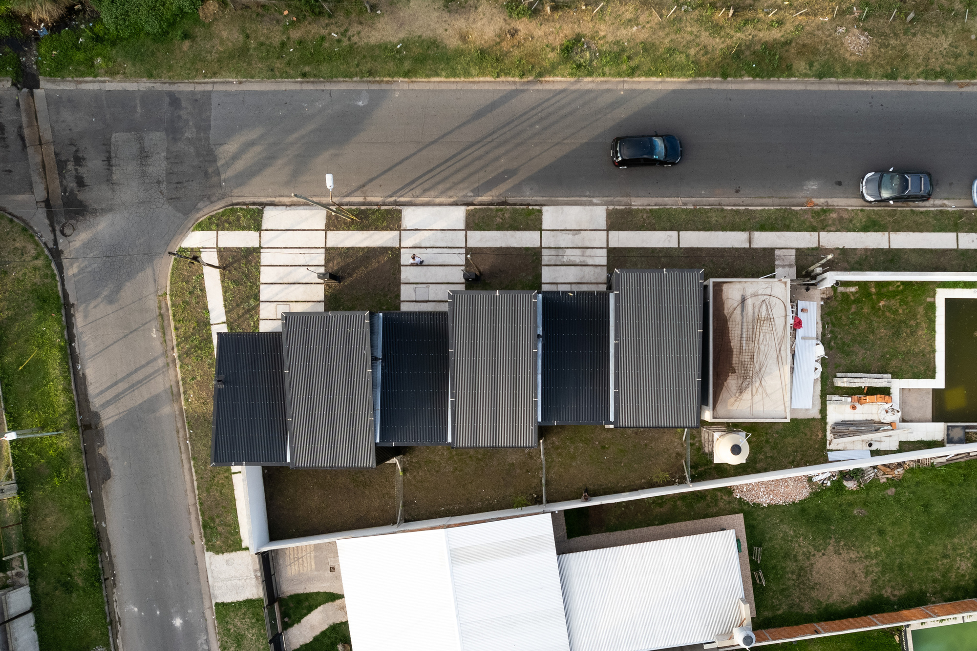 Drone view of the linear townhouse roofline with dark corrugated metal panels and courtyard voids beside the street